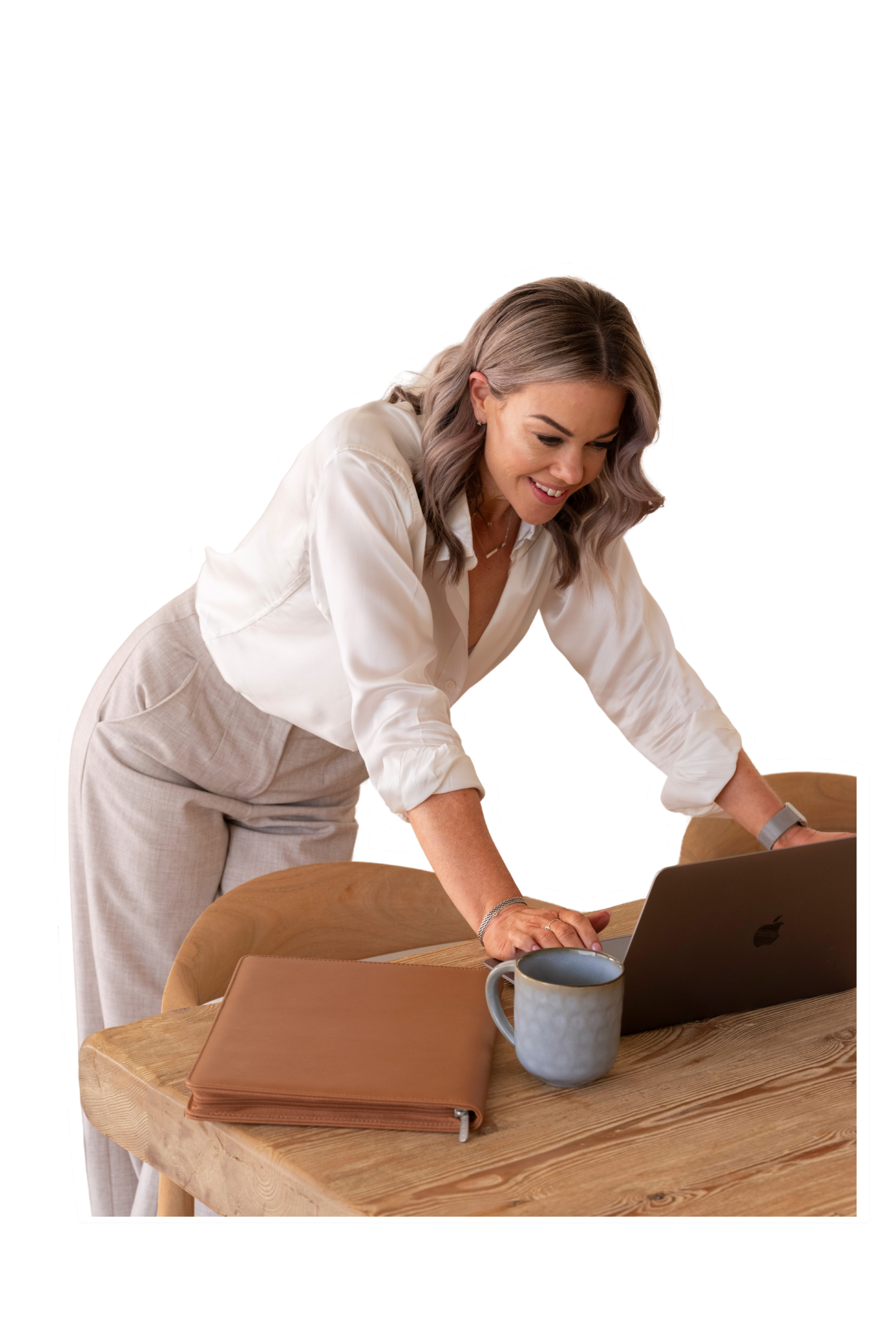A woman with shoulder-length gray hair leaning over a wooden table, looking at a laptop, smiling. On the table, there is a gray cup, a closed tan folder, and a laptop.