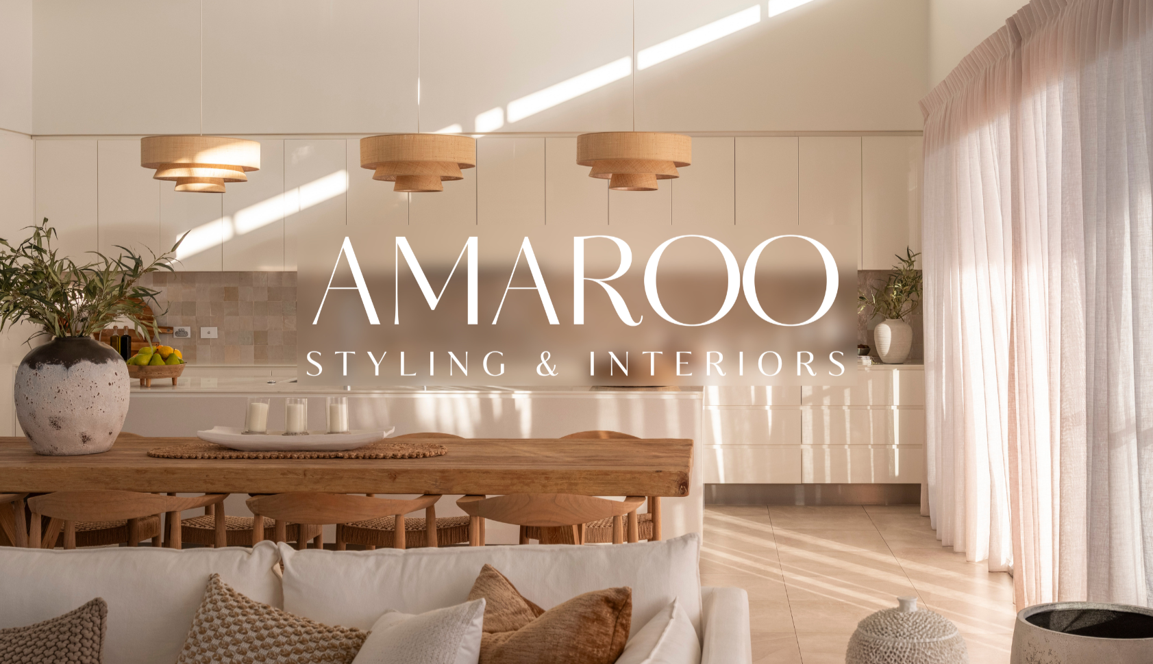 Modern kitchen and living room interior with natural light, wooden dining table, beige curtains, and decorative pillows, featuring a logo for Amaroo Styling & Interiors.