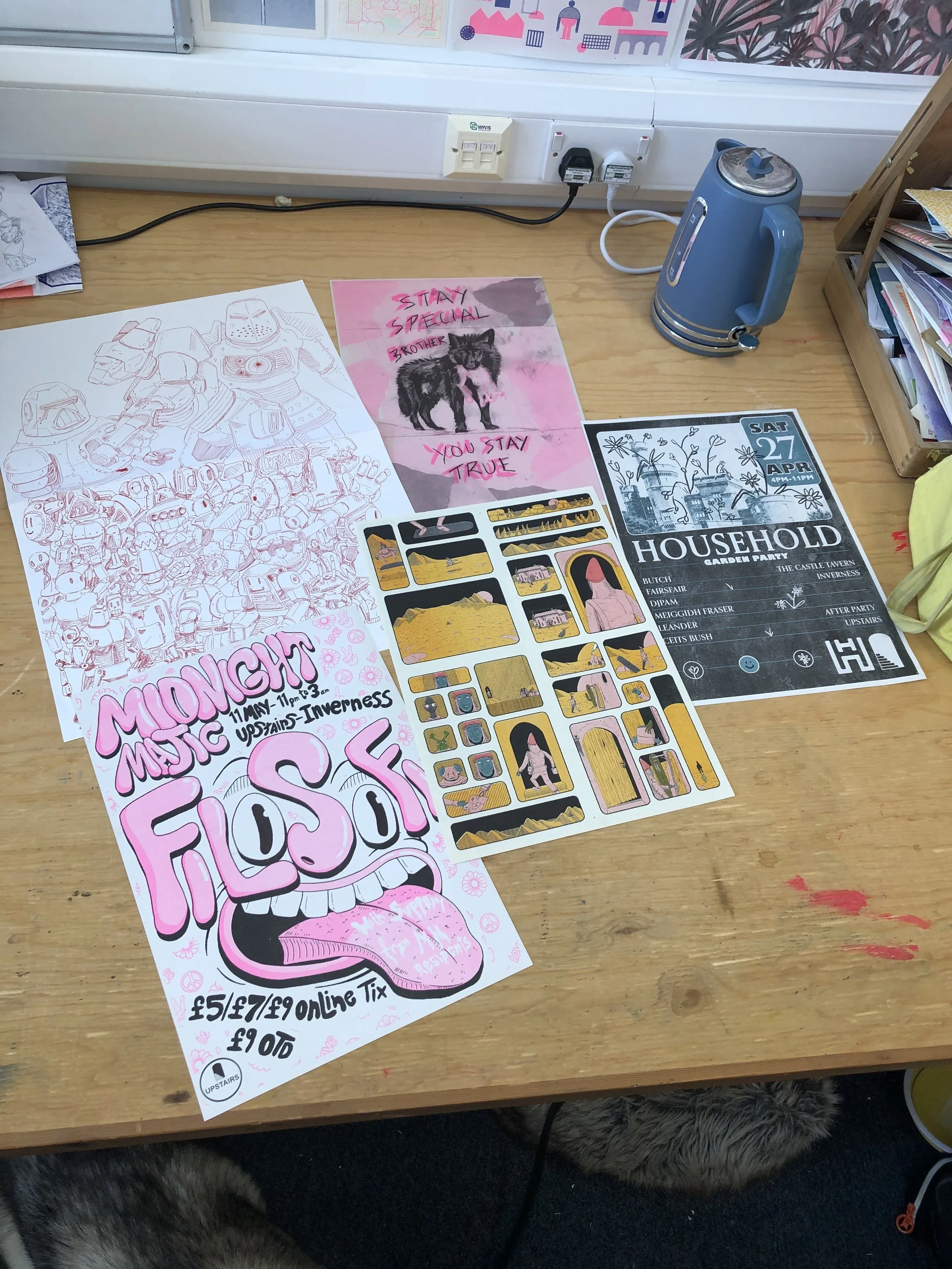Colorful posters and flyers on a wooden table, including a drawing of a robot, a pink poster with a black shaggy dog illustration and the text 'Stay Special Brother You Stay True,' a black and white poster with the date 'Sat 27 Apr,' and other various designs, in a room with a bookshelf and electric sockets.