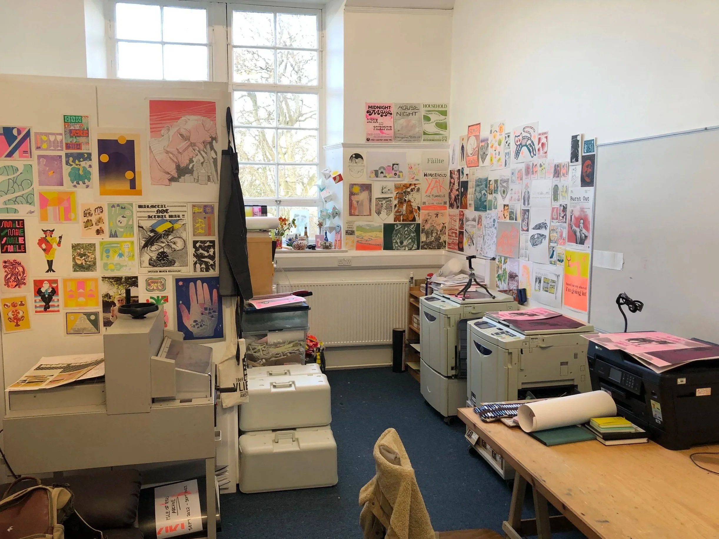 A room with white walls covered with colorful posters and artwork. There are three large windows letting in natural light. The room contains multiple photocopiers, a desk, various paper supplies, and some chairs. The floor is carpeted, and there's a small table and clutter near the photocopiers.