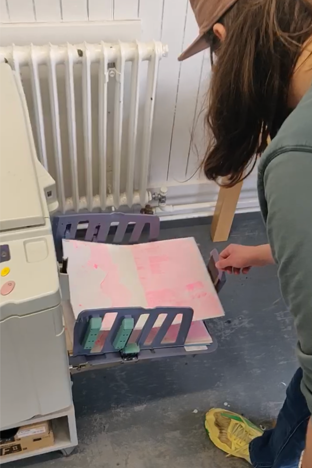 A person wearing a brown cap and yellow shoe bends over, looking at a tray holding pink documents or files in a room with a radiator on the wall and a gray floor.
