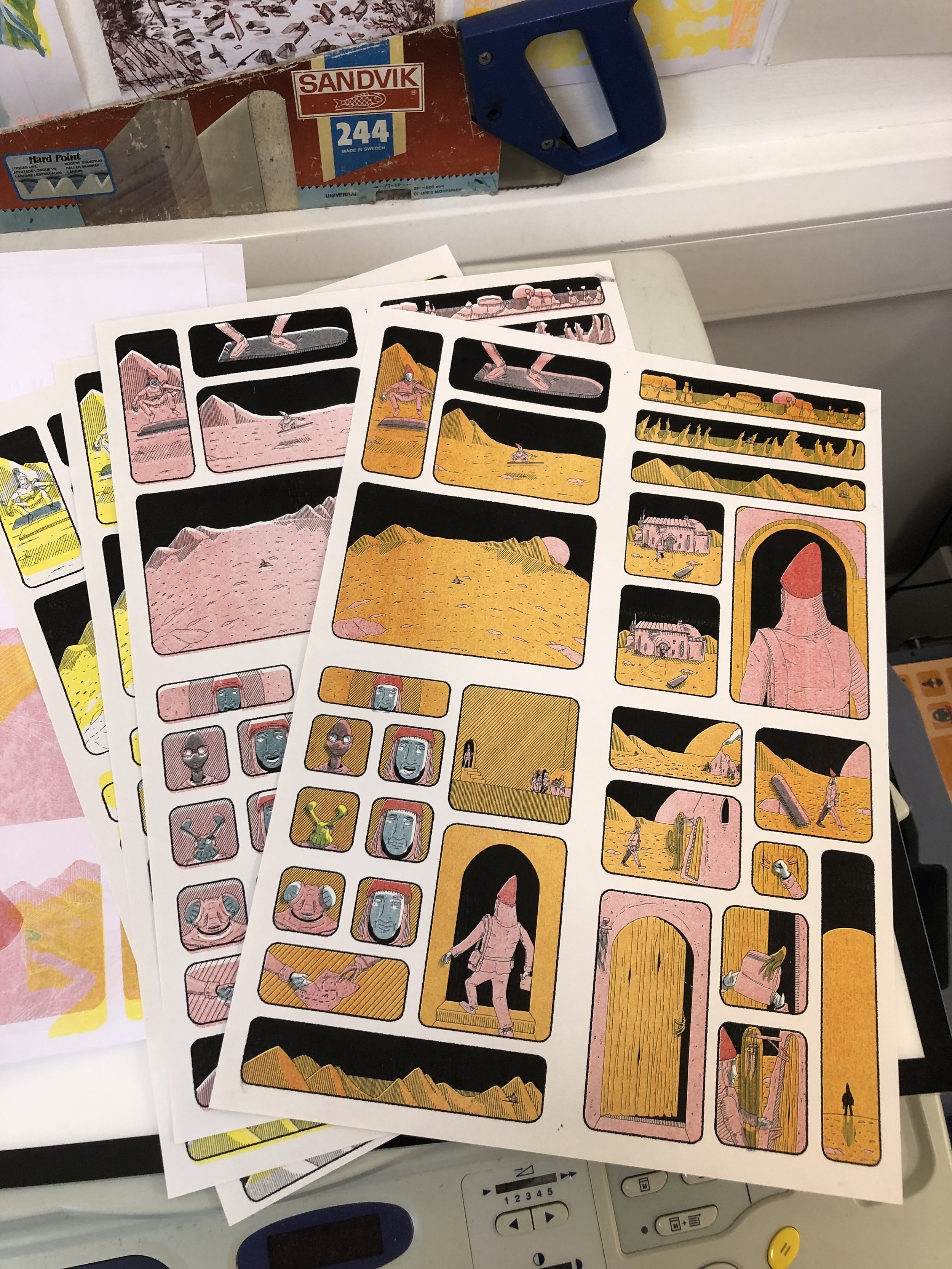 Colorful stickers on white sheets depicting desert scenes with various characters, animals, and structures, placed on a white surface with a blue and gray device in the background.