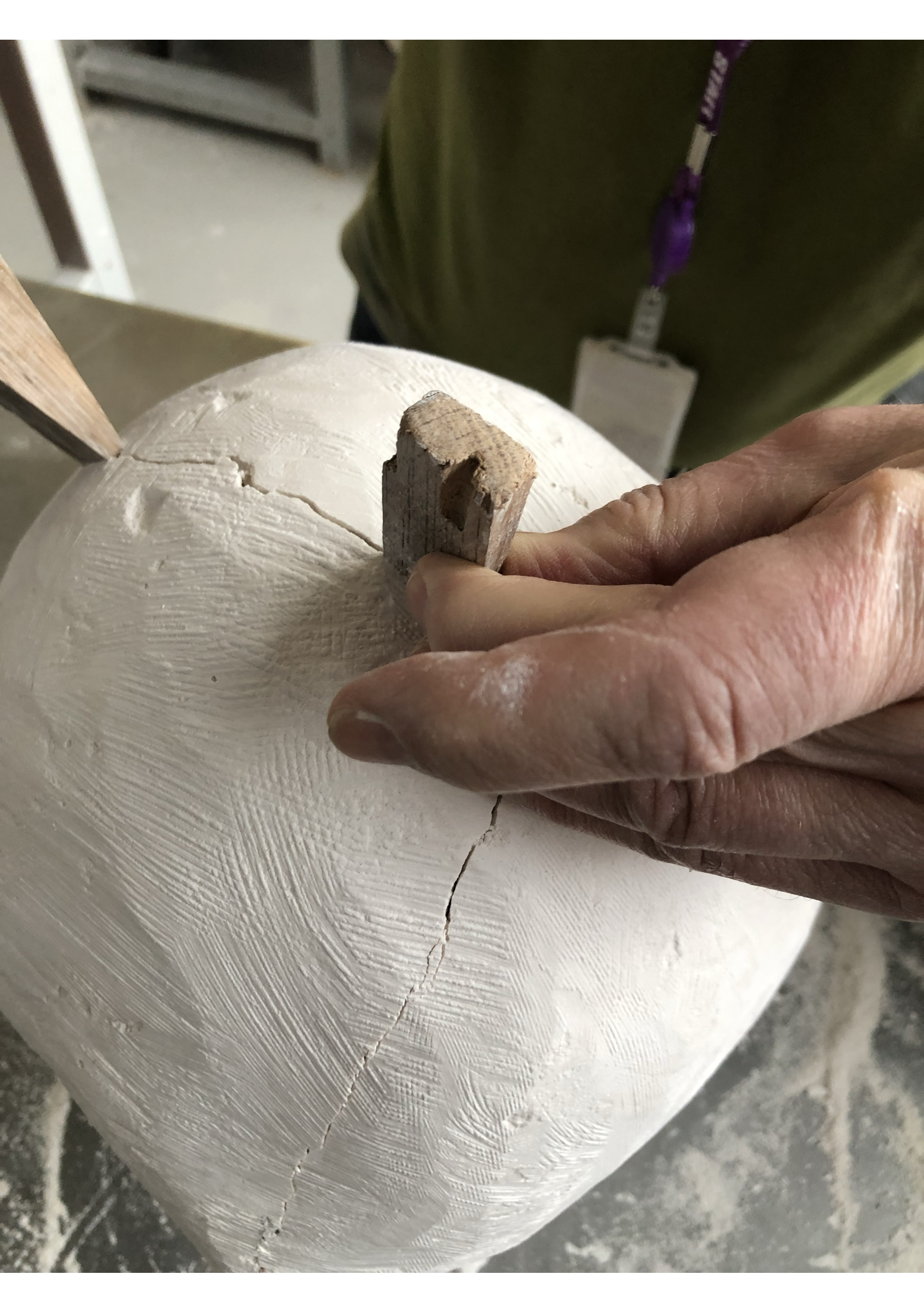 Close-up of a person's hand carving details into a round, white wooden surface with a small chisel tool, with additional woodworking tools and a worker wearing a green shirt in the background.