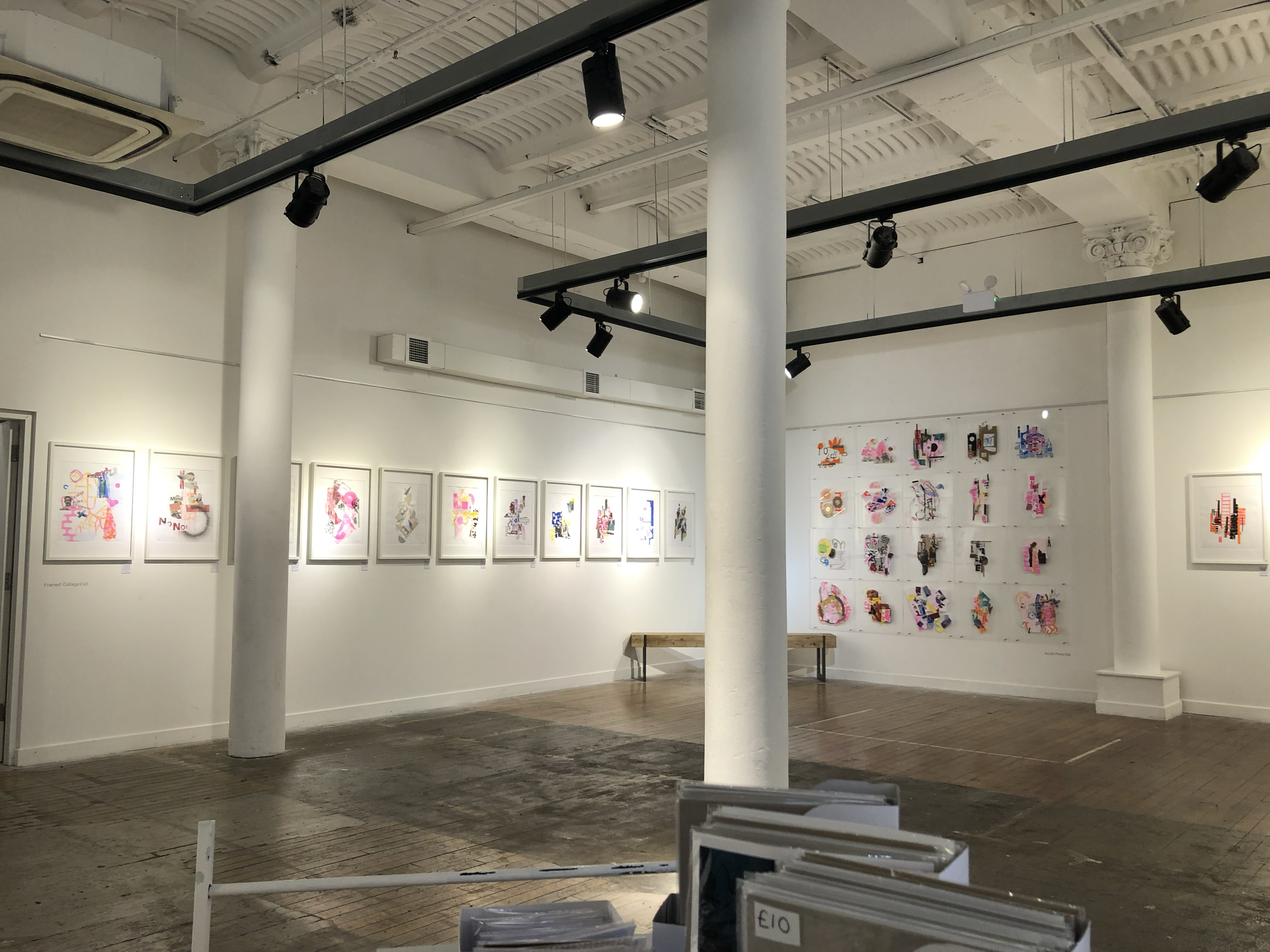 Photo of an art gallery or exhibition space with paintings displayed on the white walls, ceiling lights, and display racks in the foreground.