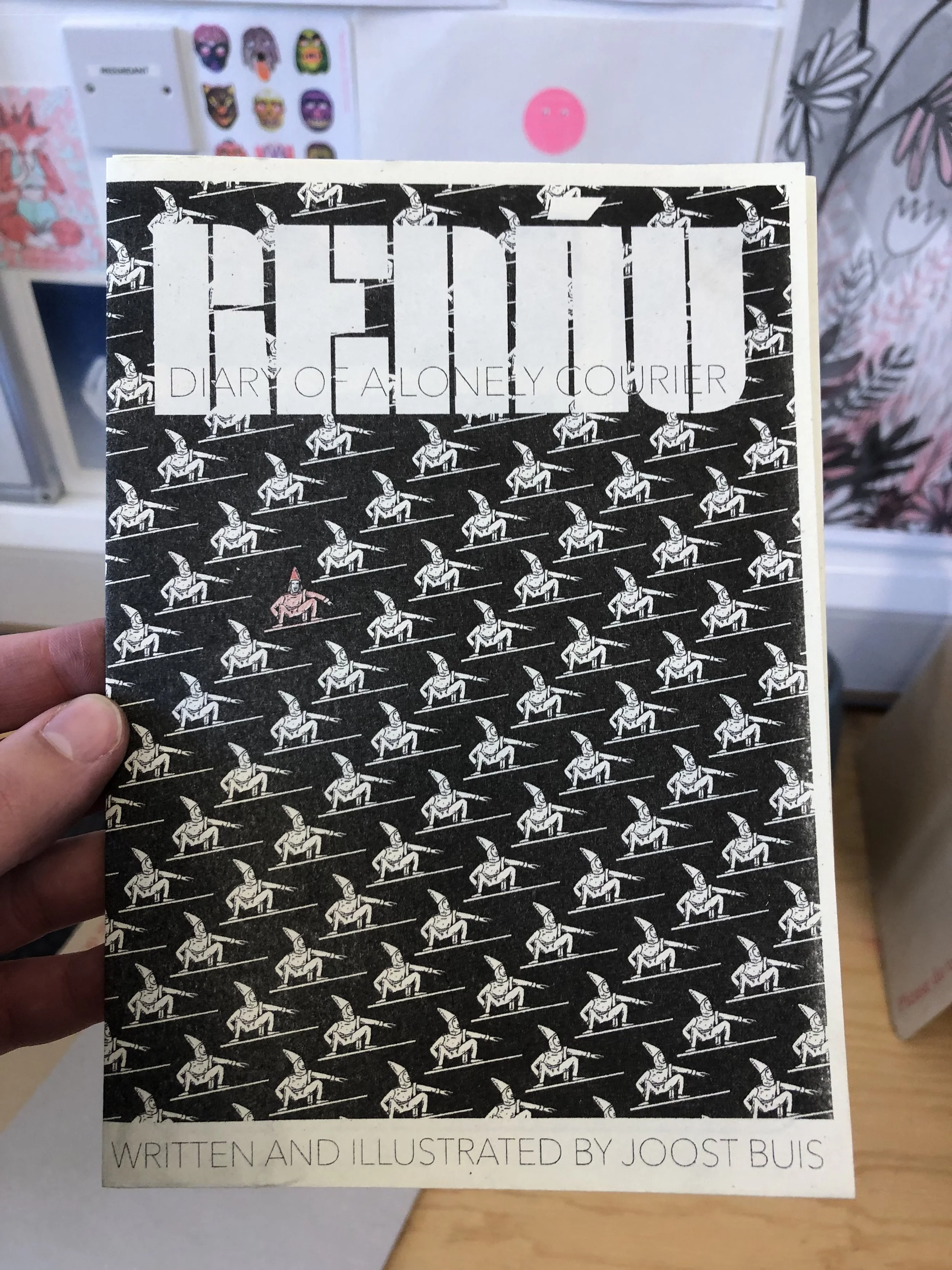 A person holding a book titled 'Renau - The dairy of a lonely courier' with illustrations of soldiers on the cover. The background includes a wall with stickers and drawings.