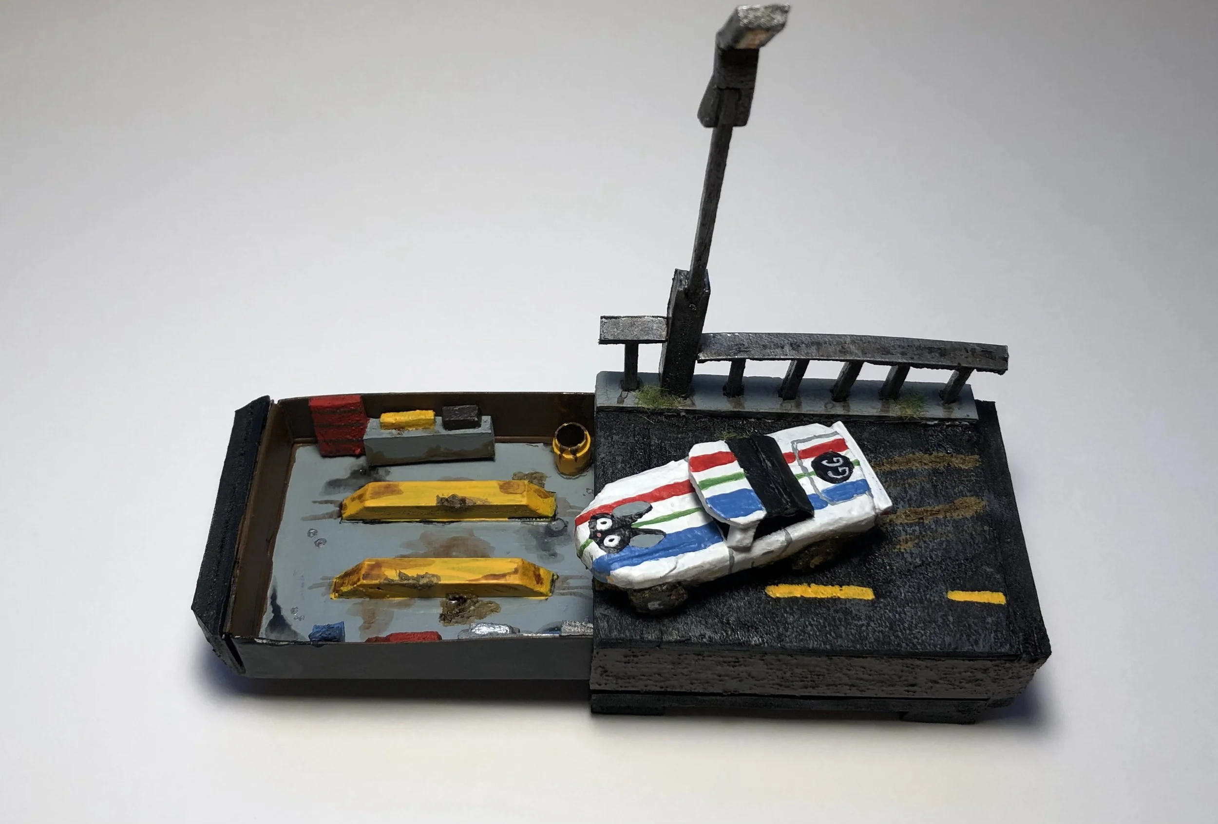 Miniature diorama of a train station with painted wooden steps, platform, and a small white crate with painted red, blue, green, and black patterns. A small black and white bird figure stands atop a wrapped package near the platform.