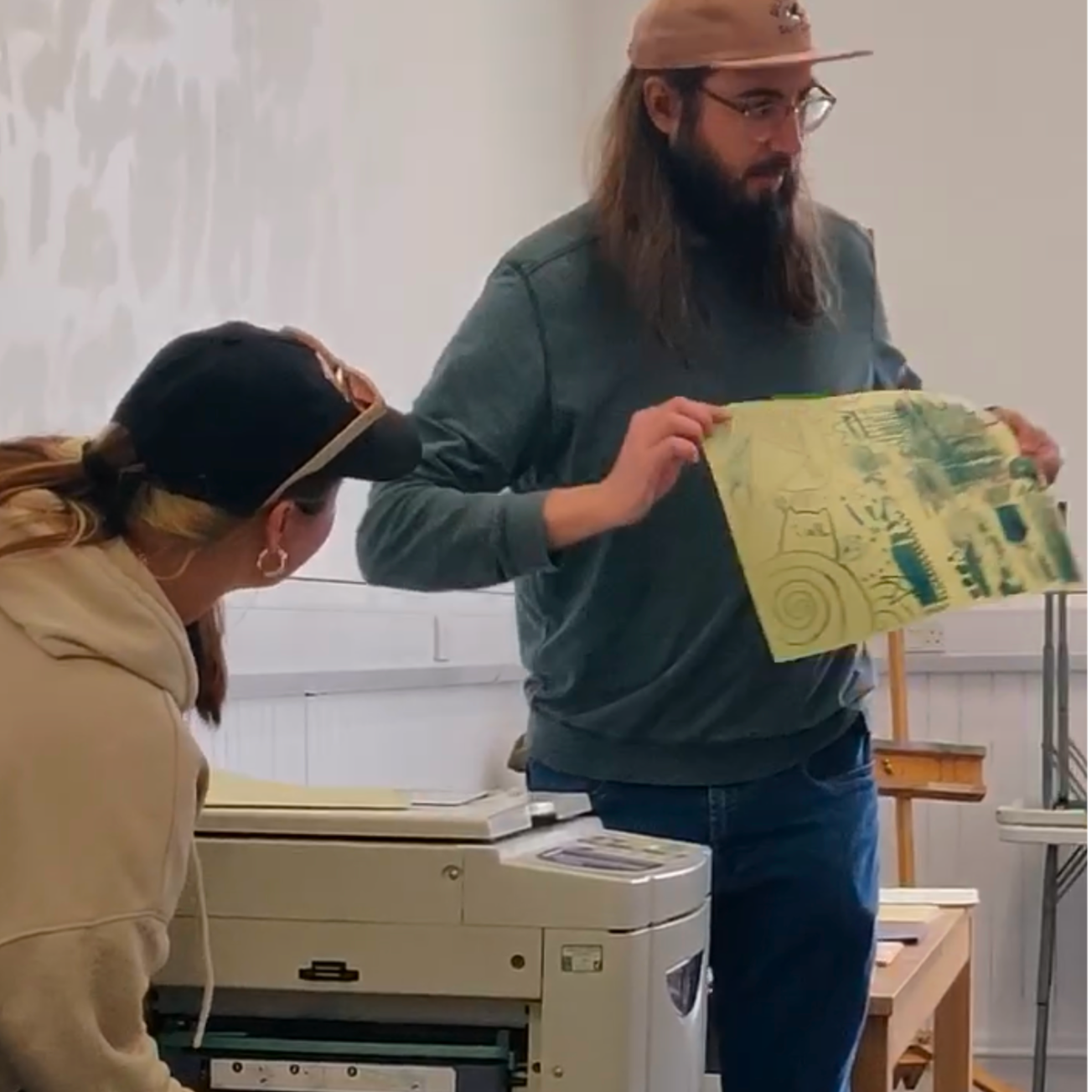 A man with long hair and a beard, wearing glasses and a pink cap, is holding a colorful piece of artwork or poster. A woman in a beige hoodie and black cap is sitting nearby, looking at the artwork. There is a photocopier or printer in front of them and a wooden easel in the background.