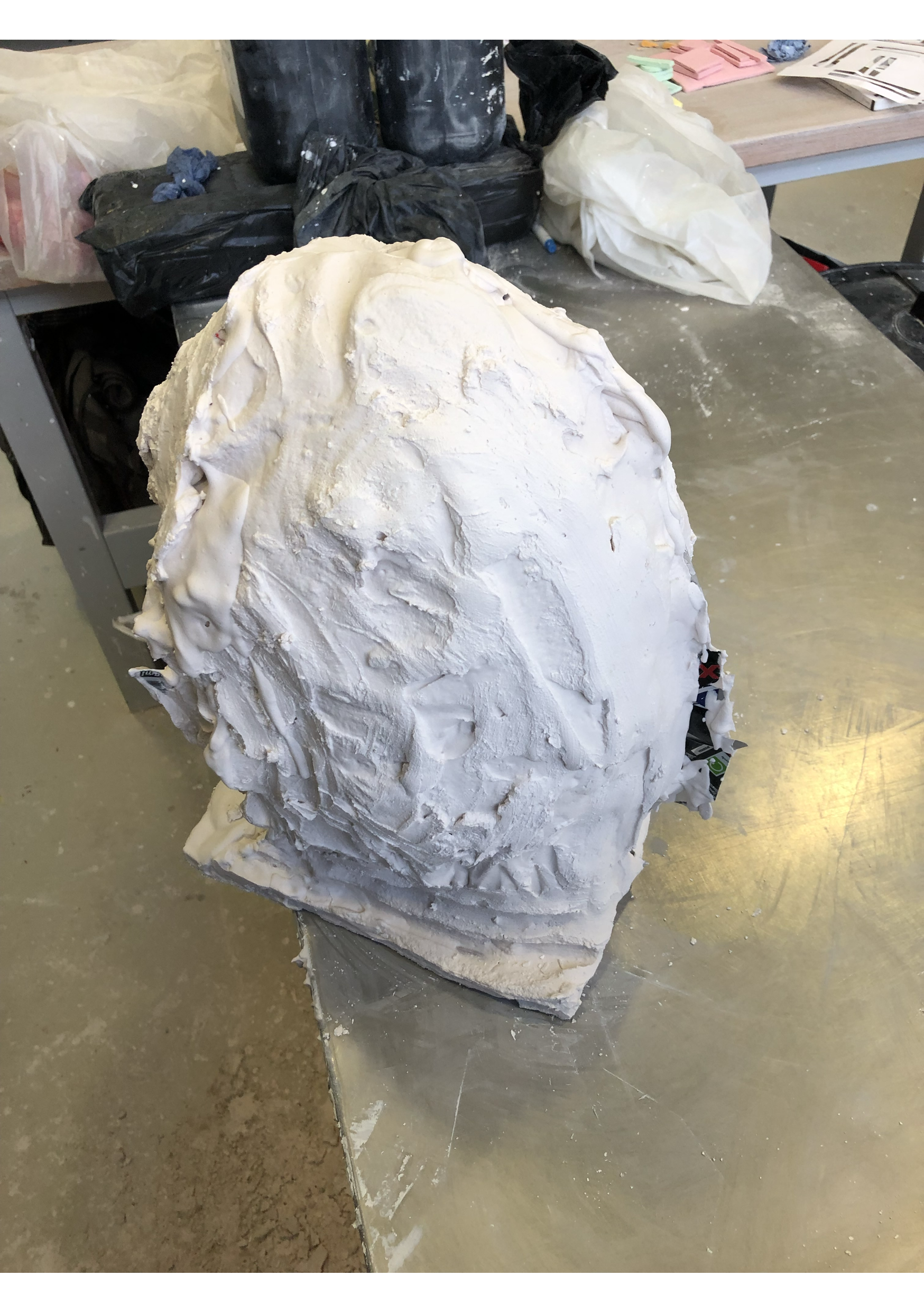 A workshop table with a large white plaster sculpture or mold and various bags and supplies surrounding it.