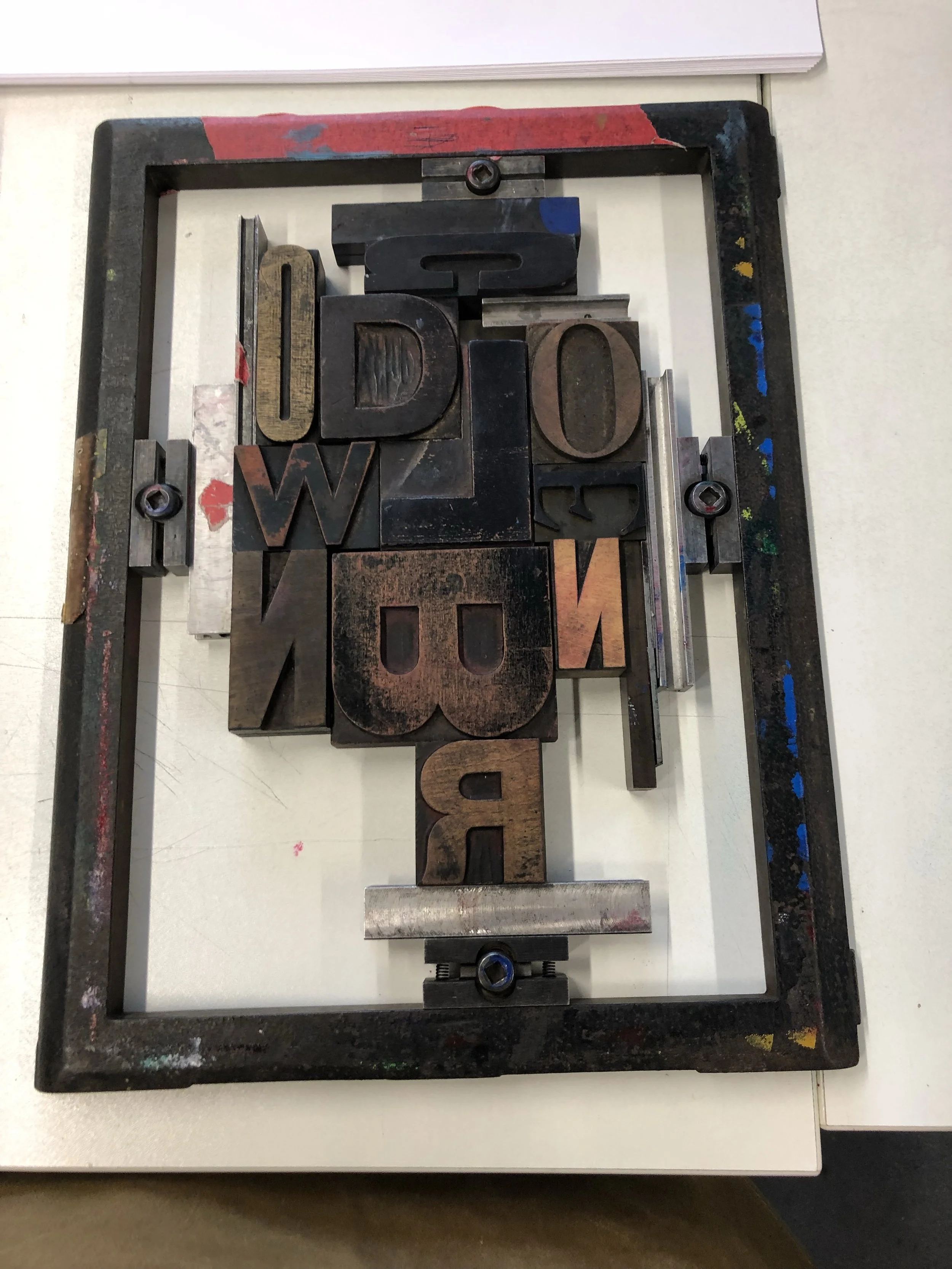 Vintage letterpress printing presses with metal blocks spelling out words, arranged on a metal frame with paint splatters, seen from above on a white surface.