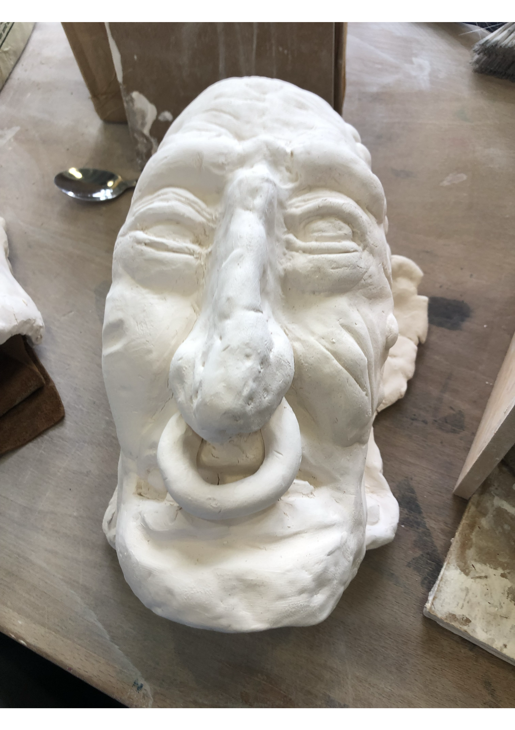 A white sculpted mask resembling an elderly person's face with detailed wrinkles and features, mounted on a base, placed on a wooden surface.