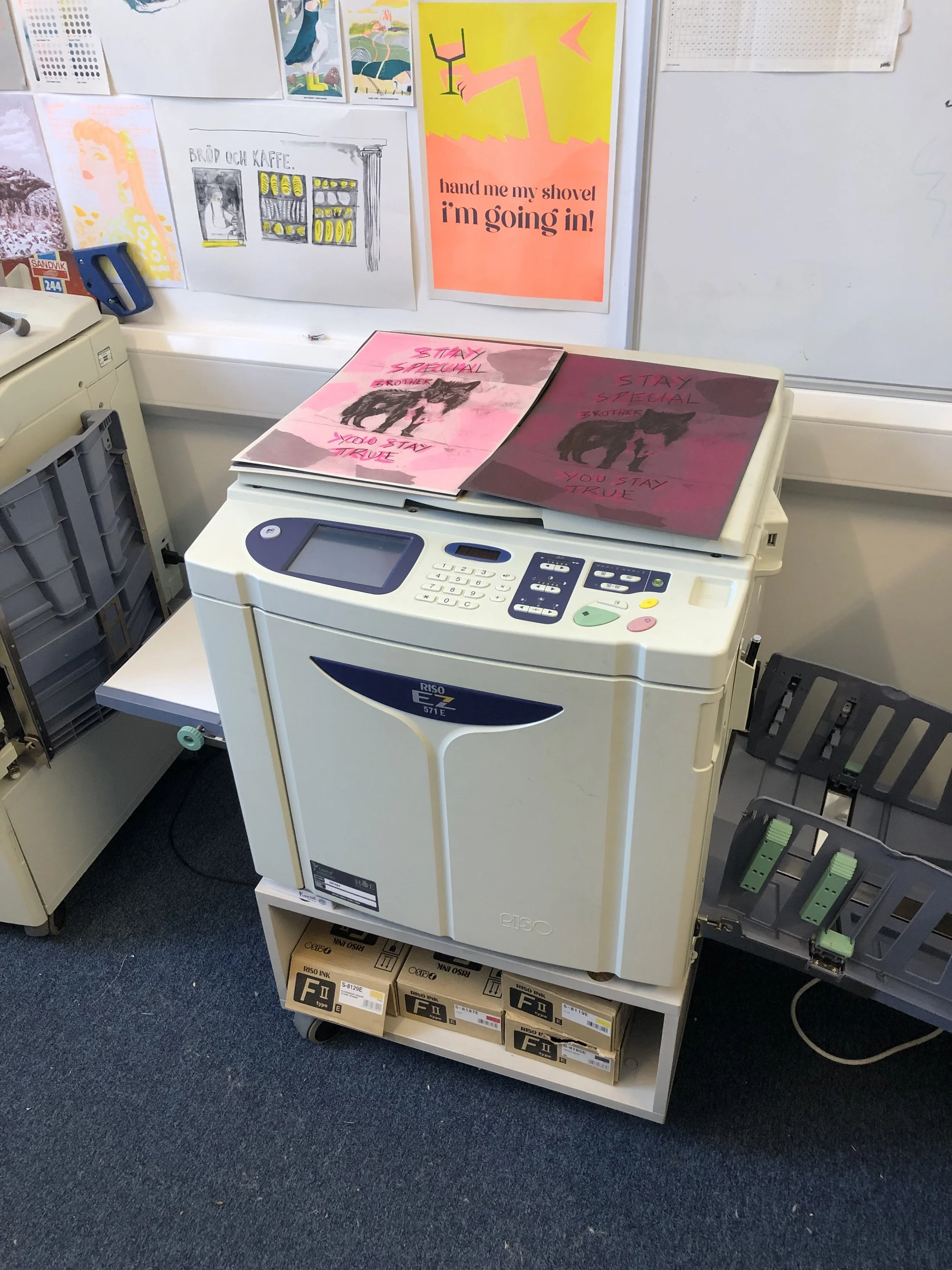RISO printer on a white cart with boxes of ink below it, surrounded by various posters and drawings on the wall.