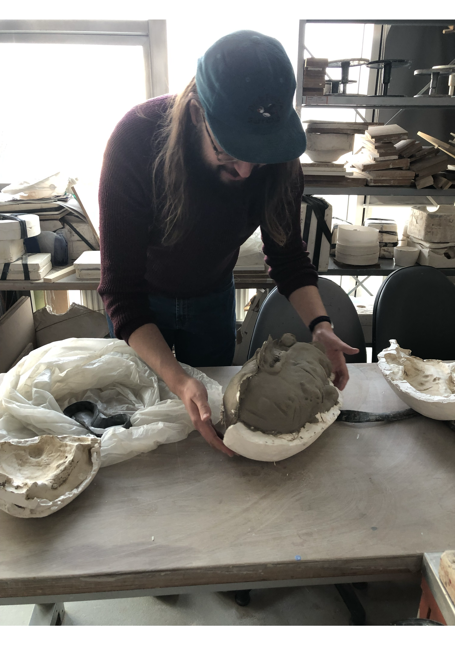 A person wearing a blue baseball cap and glasses is working on a mold of a face sculpture, surrounded by plaster and clay on a large table in an art studio with shelves of supplies and windows in the background.