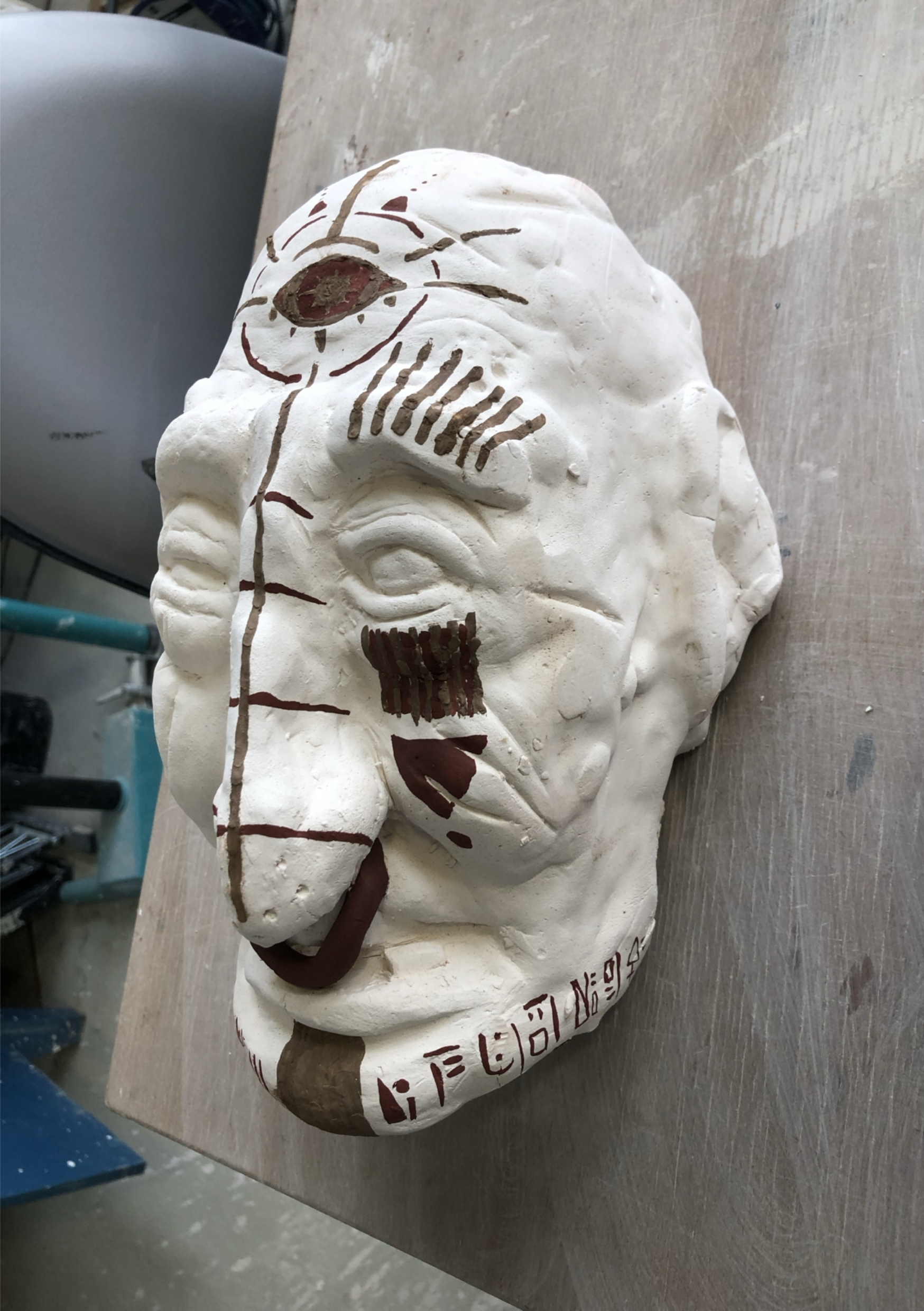 A white skull sculpture with painted facial features and red markings. It is lying on a wooden surface in a workshop or storage area.