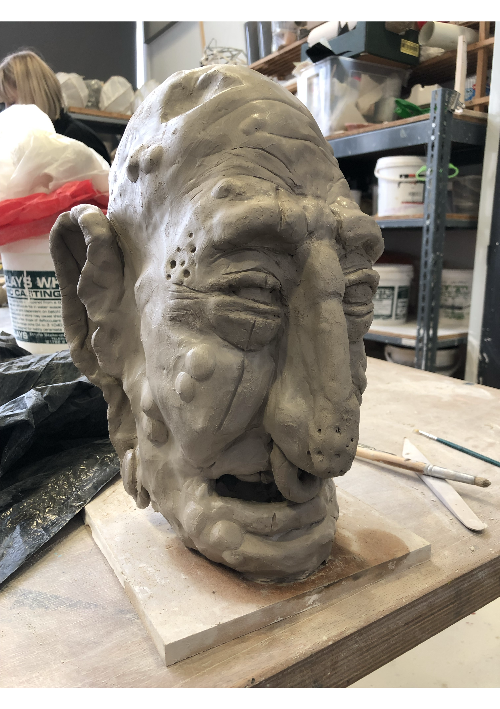 Clay sculpture of a face with expressive features, placed on a wooden workbench in an artist's studio surrounded by art supplies and shelves.
