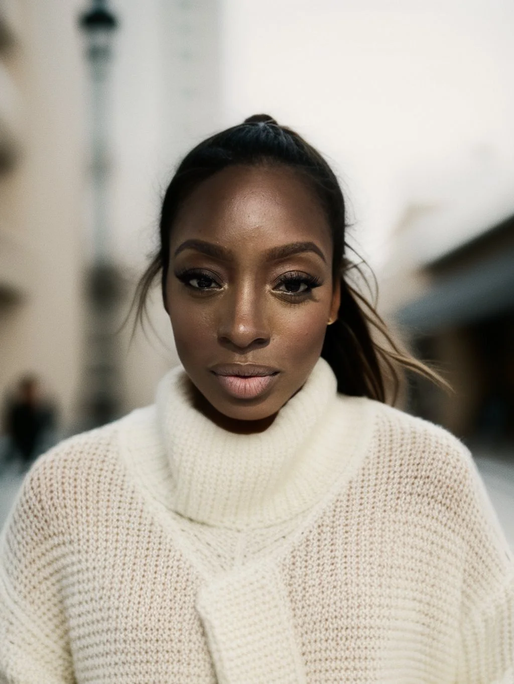 Eunice Adebiyi, brown skin, dark makeup, and a high ponytail wearing a cream-colored, chunky-knit turtleneck sweater, standing outdoors with blurred buildings in the background.
