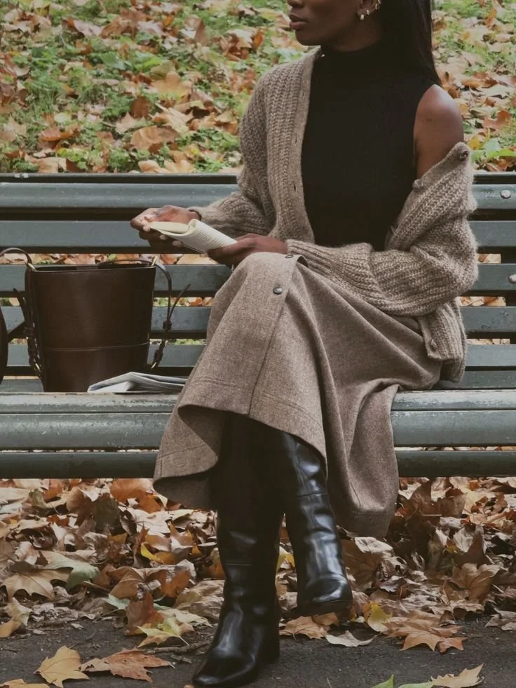 A woman sitting on a park bench outdoors among autumn leaves, reading a book, dressed in a black top, chunky cardigan, plaid skirt, and knee-high black boots.