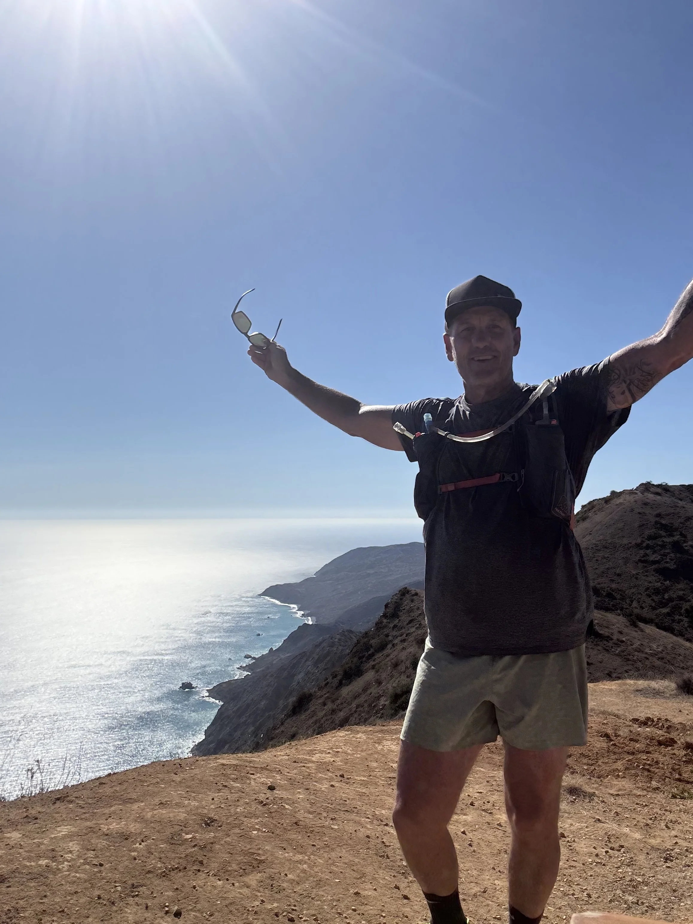 A man smiling with open arms on a hilltop overlooking the ocean, holding sunglasses in one hand, wearing a black t-shirt, tan shorts, and a hat, with a backpack and a hydration tube.