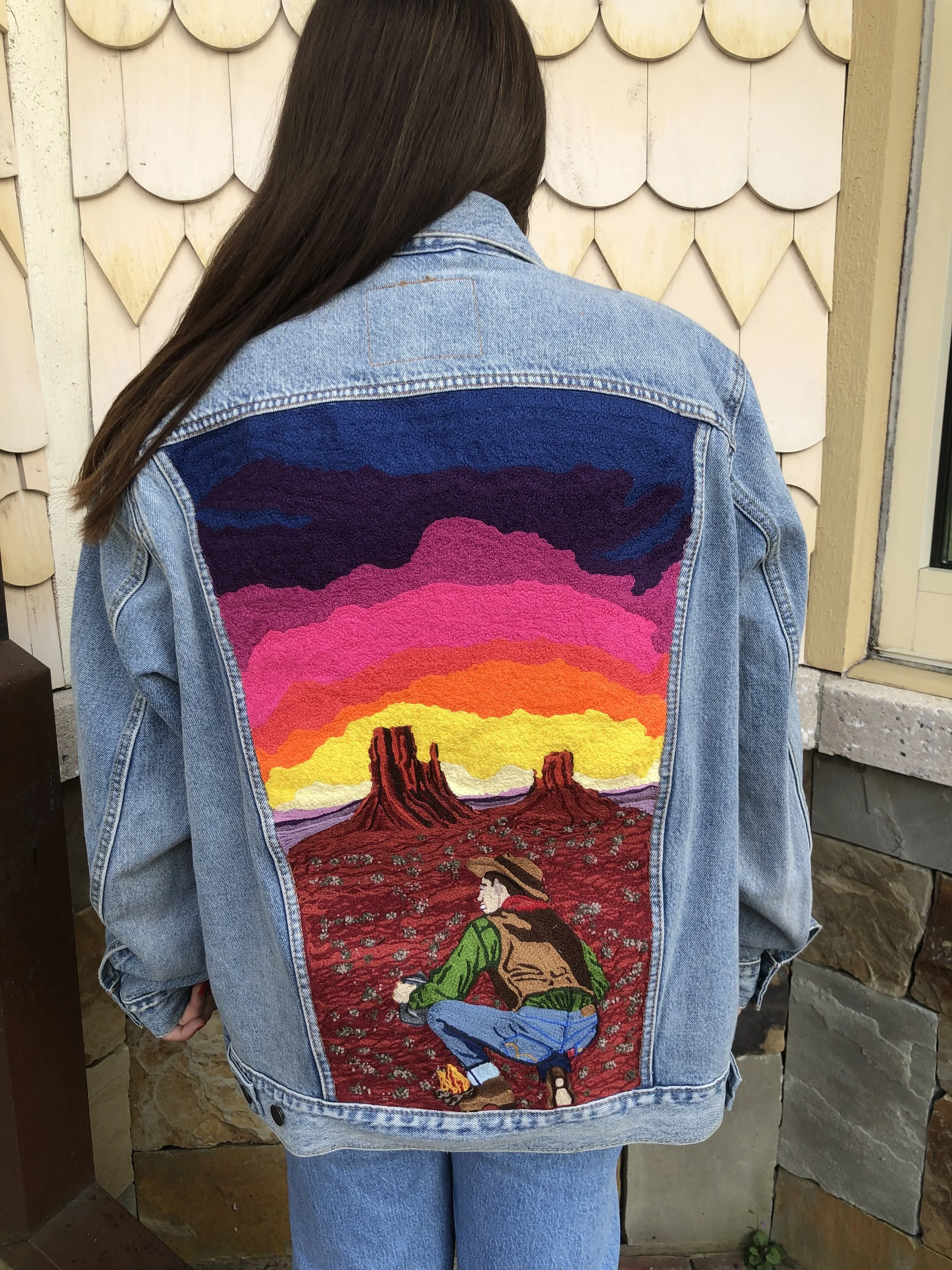 A person wearing a denim jacket with a large, colorful embroidered scene on the back depicting a desert landscape with two large rock formations, a sunset sky with vibrant pink, purple, orange, and yellow hues, and a person sitting in the foreground wearing a straw hat and green shirt.