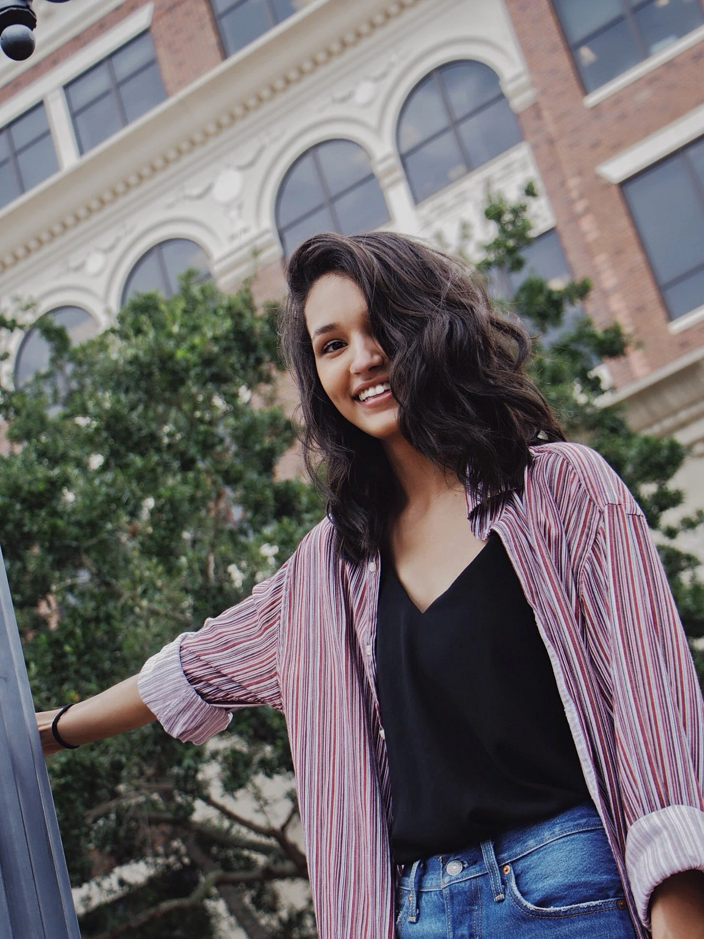 A young woman with curly dark hair smiling outdoors, standing in front of a building with arched windows and trees.