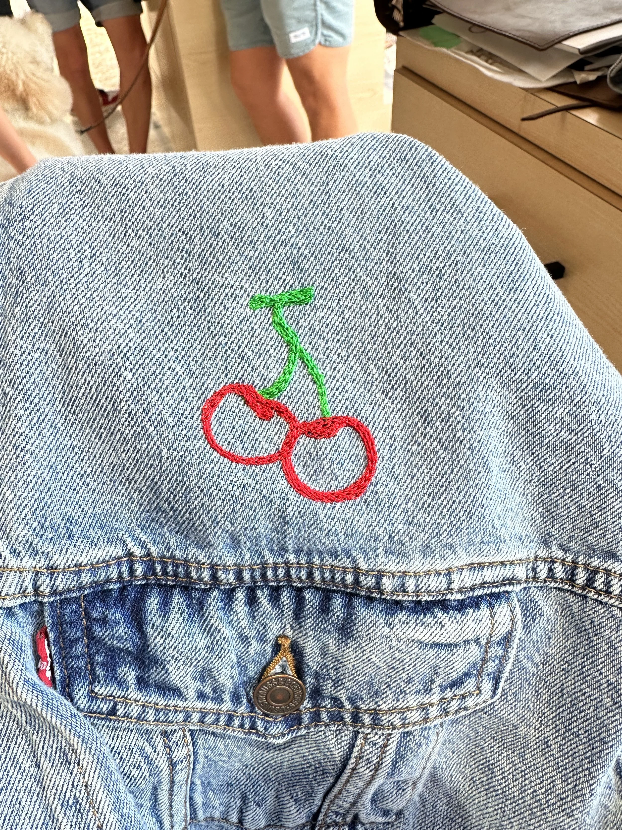 Close-up of a light blue denim jacket with a cherry embroidery in red and green thread on the back pocket.