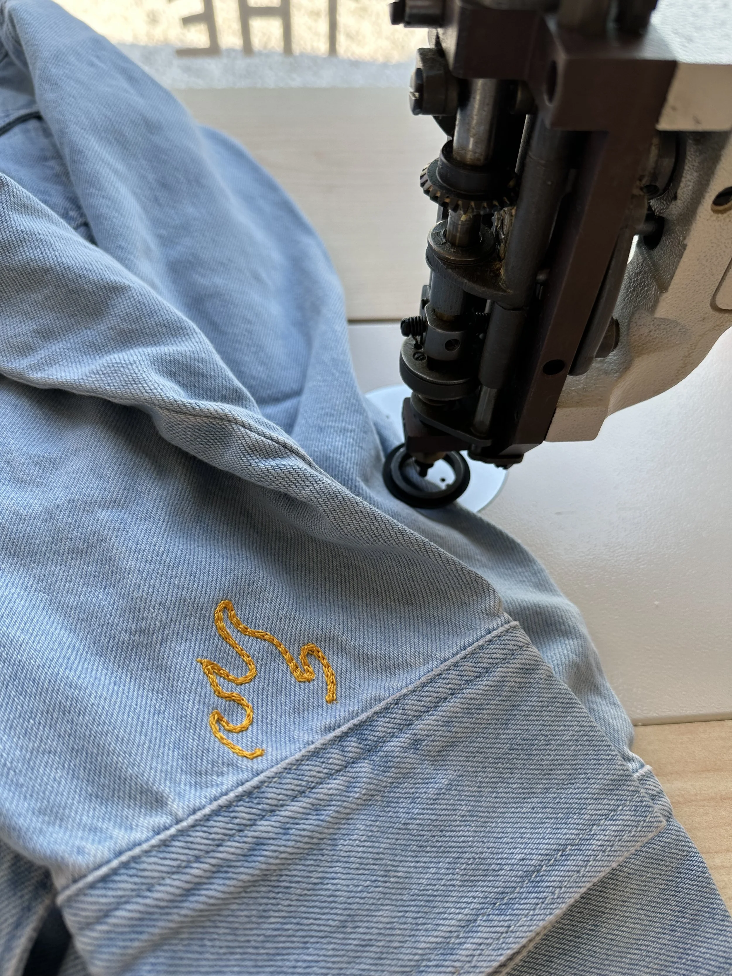 Close-up of a sewing machine stitching an embroidered yellow flame onto light blue denim fabric.