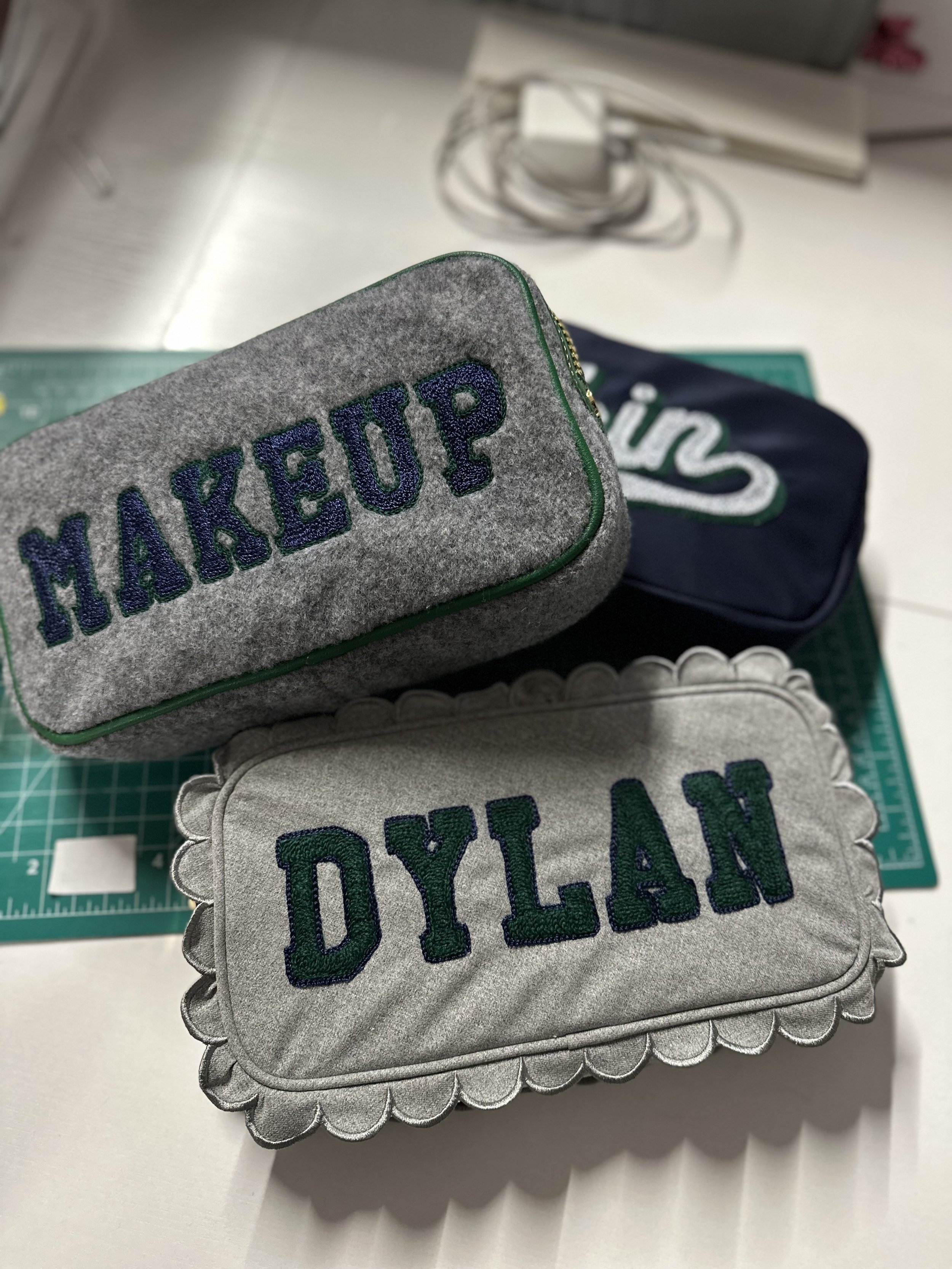 Three personalized fabric cases with embroidered names 'MAKEUP', 'DYLAN', and an unknown name in the background, placed on a green cutting mat.