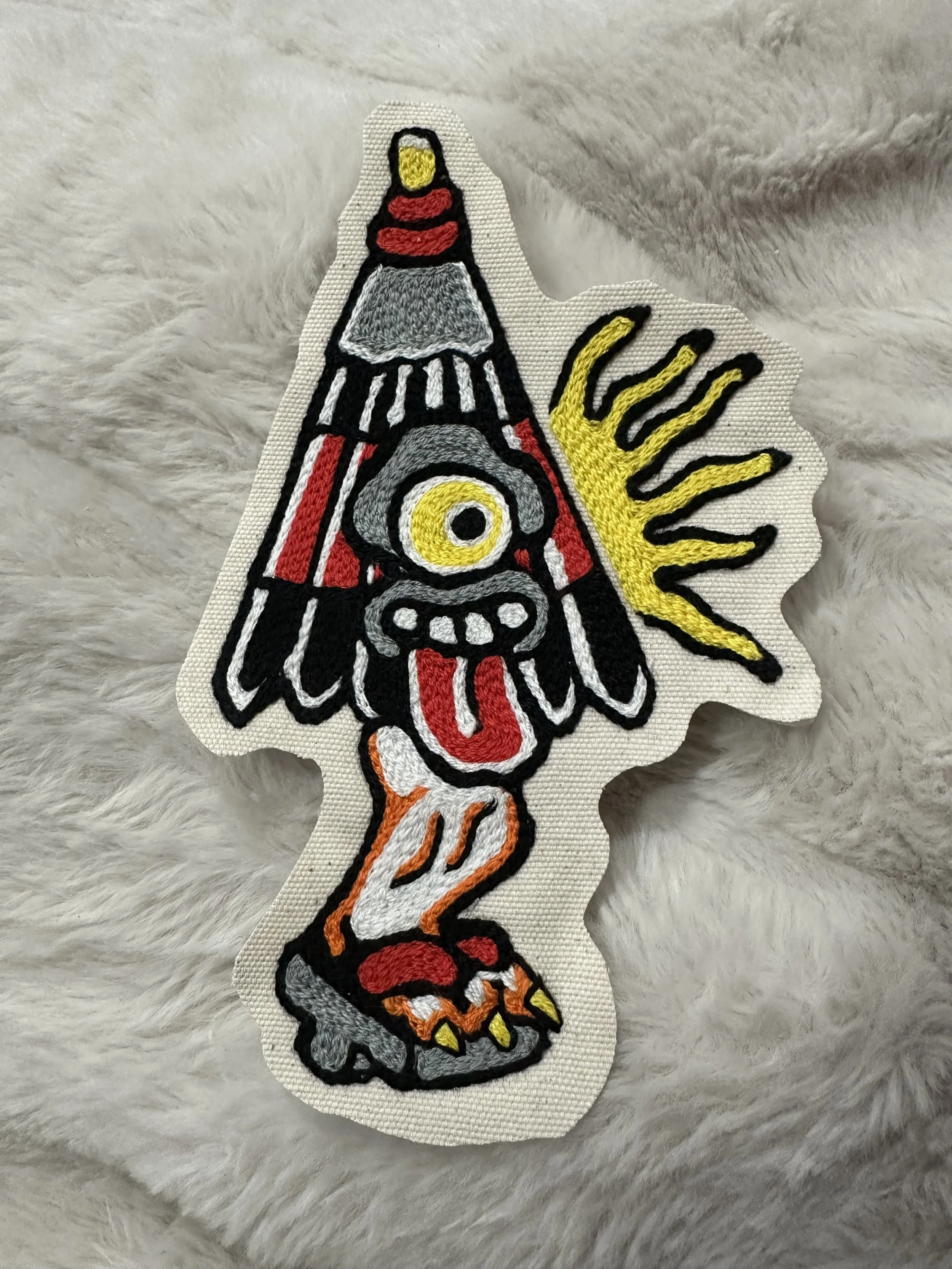 Colorful embroidered patch of a cartoonish totem pole with a bird, eye, tongue, and yellow sun rays on a soft, gray, furry surface.