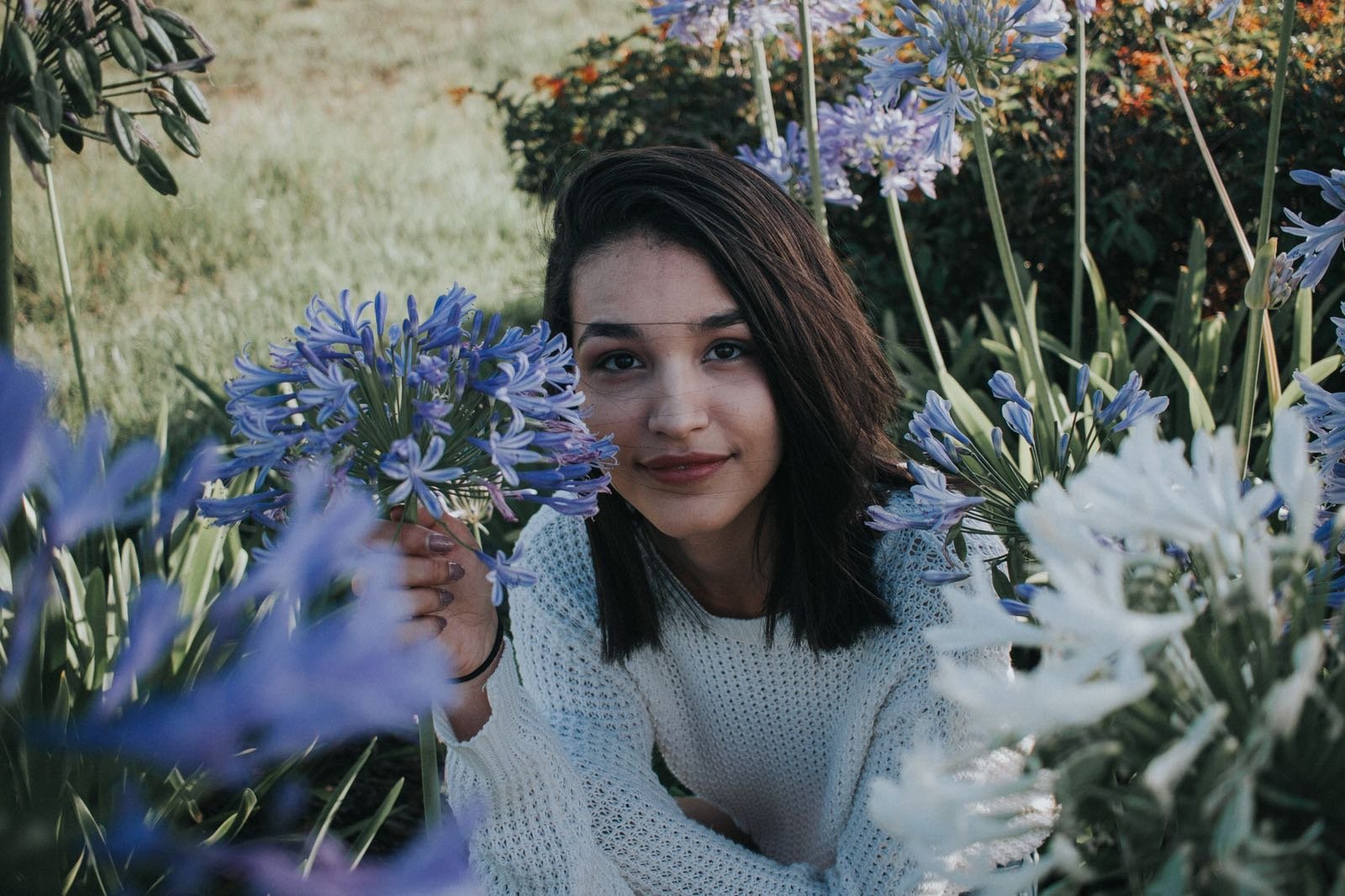 A young woman with dark hair and a white sweater is smiling while lying among purple and white flowers outdoors.