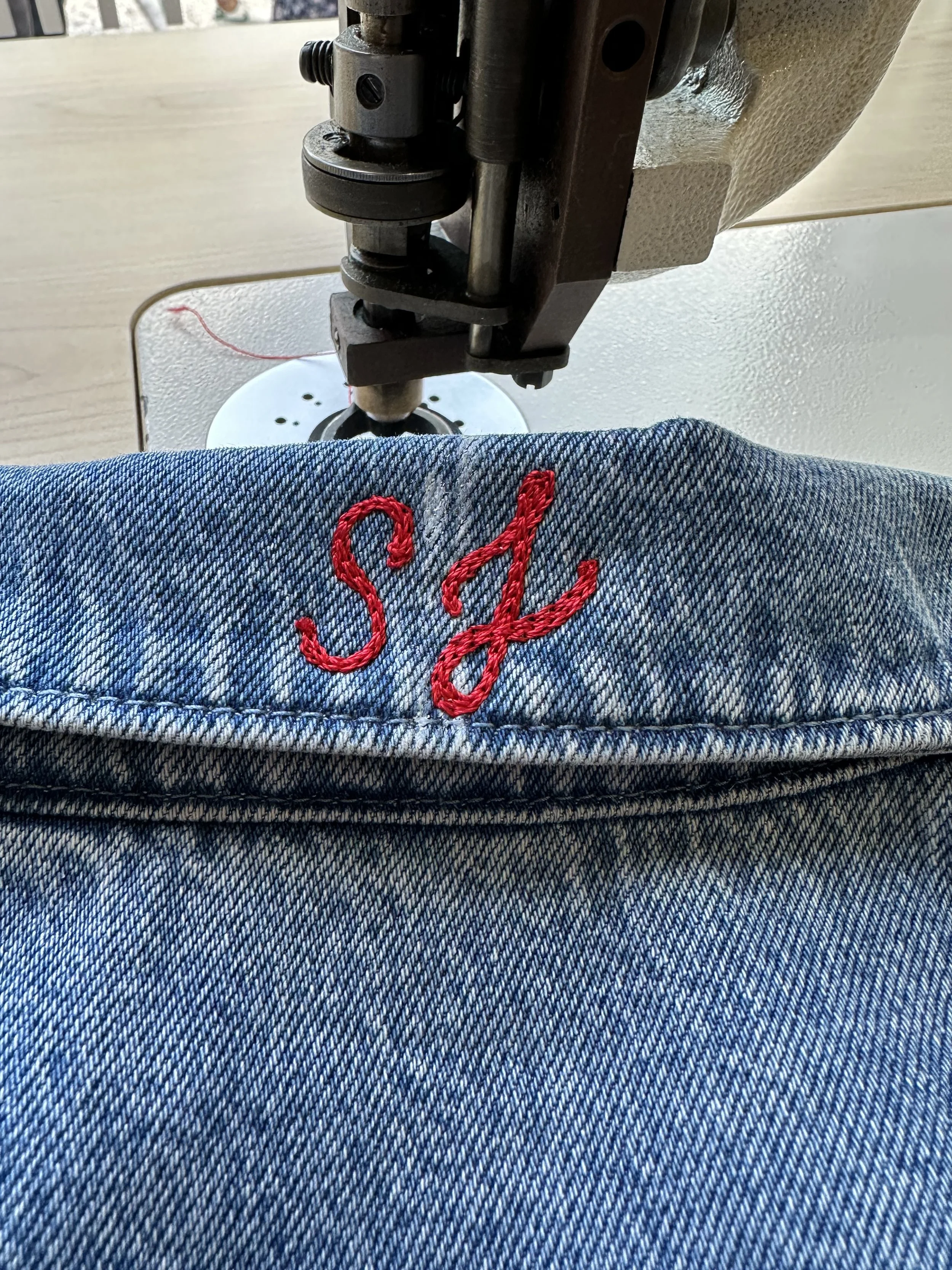 Close-up of a sewing machine stitching an embroidered red letter 'S' and 'J' on denim fabric.