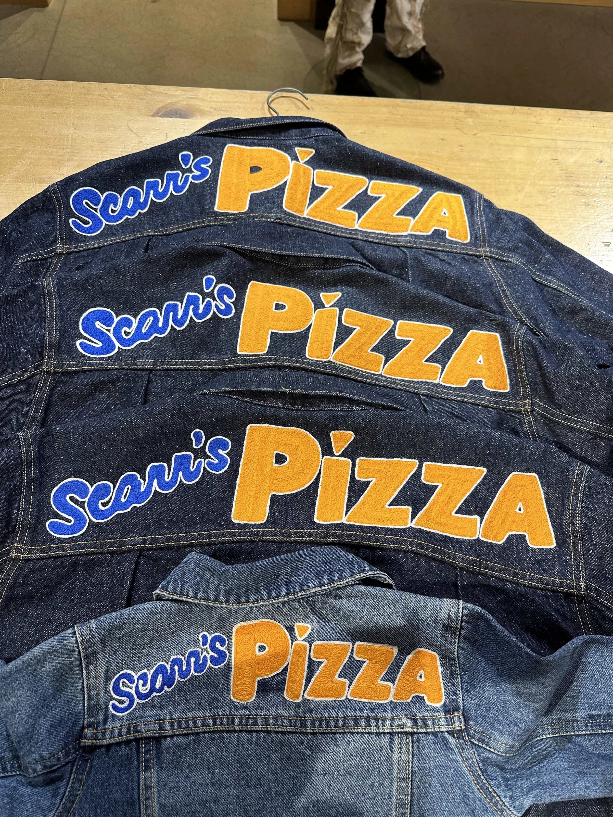 Four denim jackets with embroidered text that reads 'Scarrs's PIZZA' in blue and orange, arranged on a wooden surface.