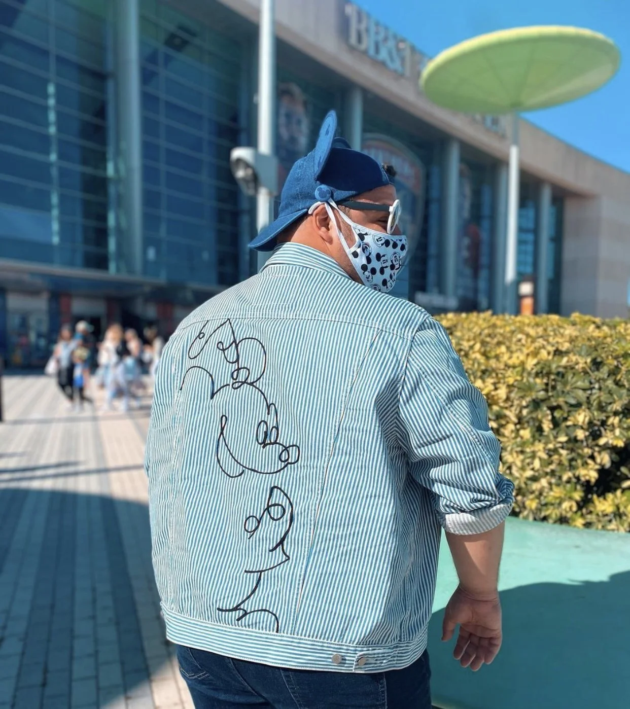 A person with a blue cap, sunglasses, and a face mask with a panda design, wearing a striped jacket with a black doodle on the back, standing outside near a large building with a green structure overhead, with people in the background.
