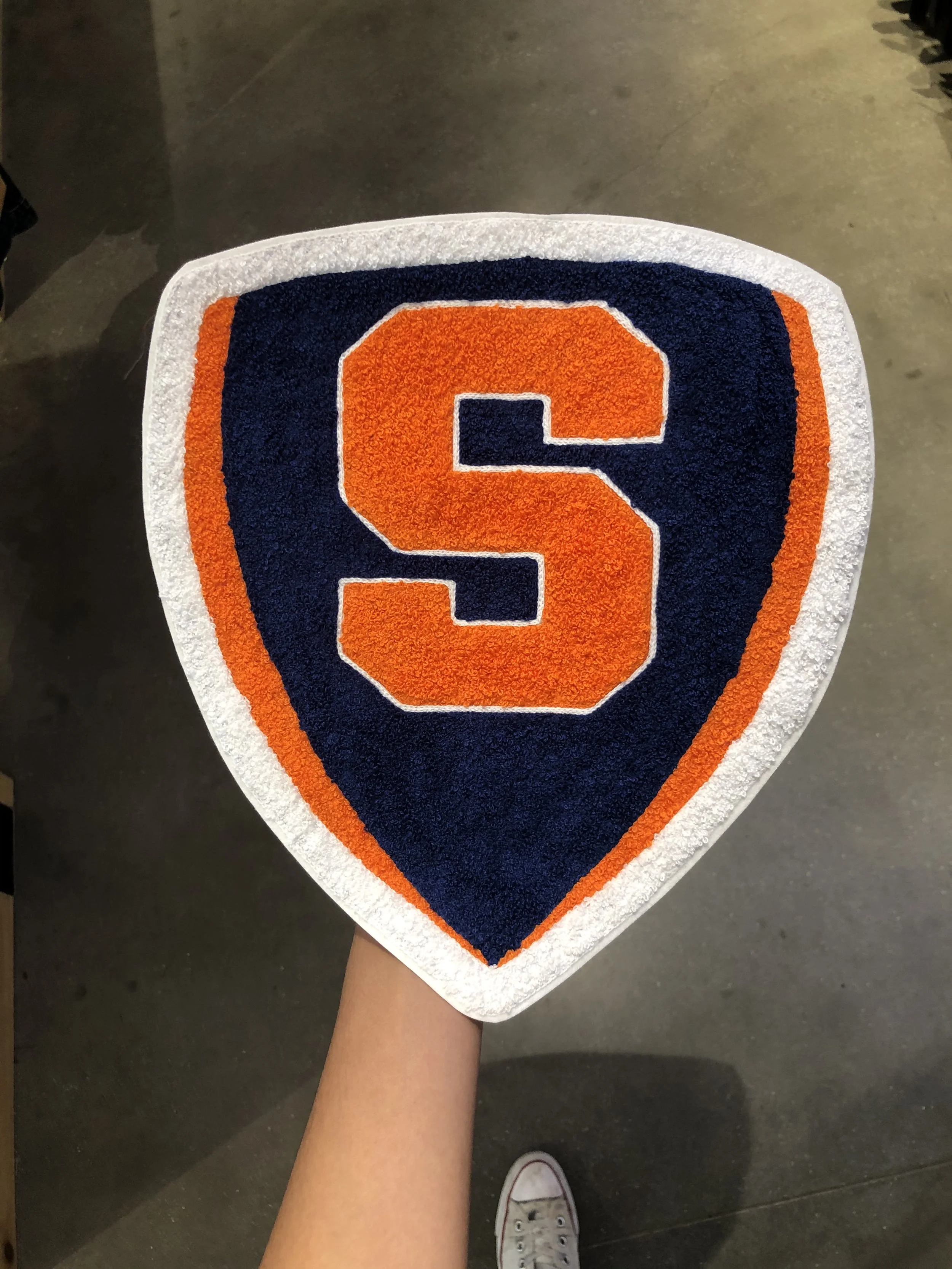 A hand holding a Syracuse University logo-shaped rug with an orange 'S' on a navy blue background, outlined with white and orange borders.