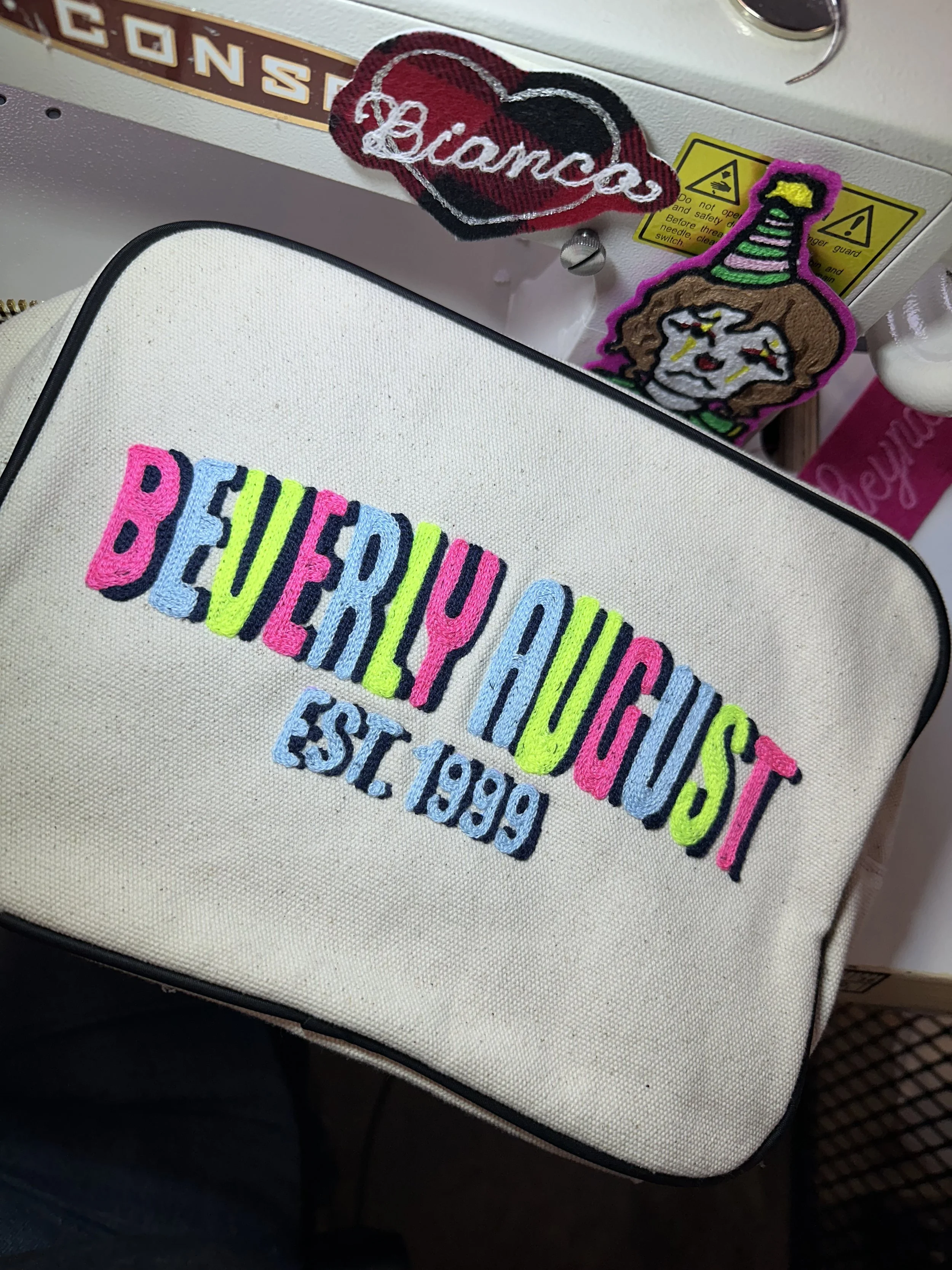 Embroidery on a white bag with colorful text saying "Beverly August" and "Est. 1999". Behind the bag are two patches, one heart-shaped with the name "Bianca" and the other with a cartoon clown face wearing a striped party hat.