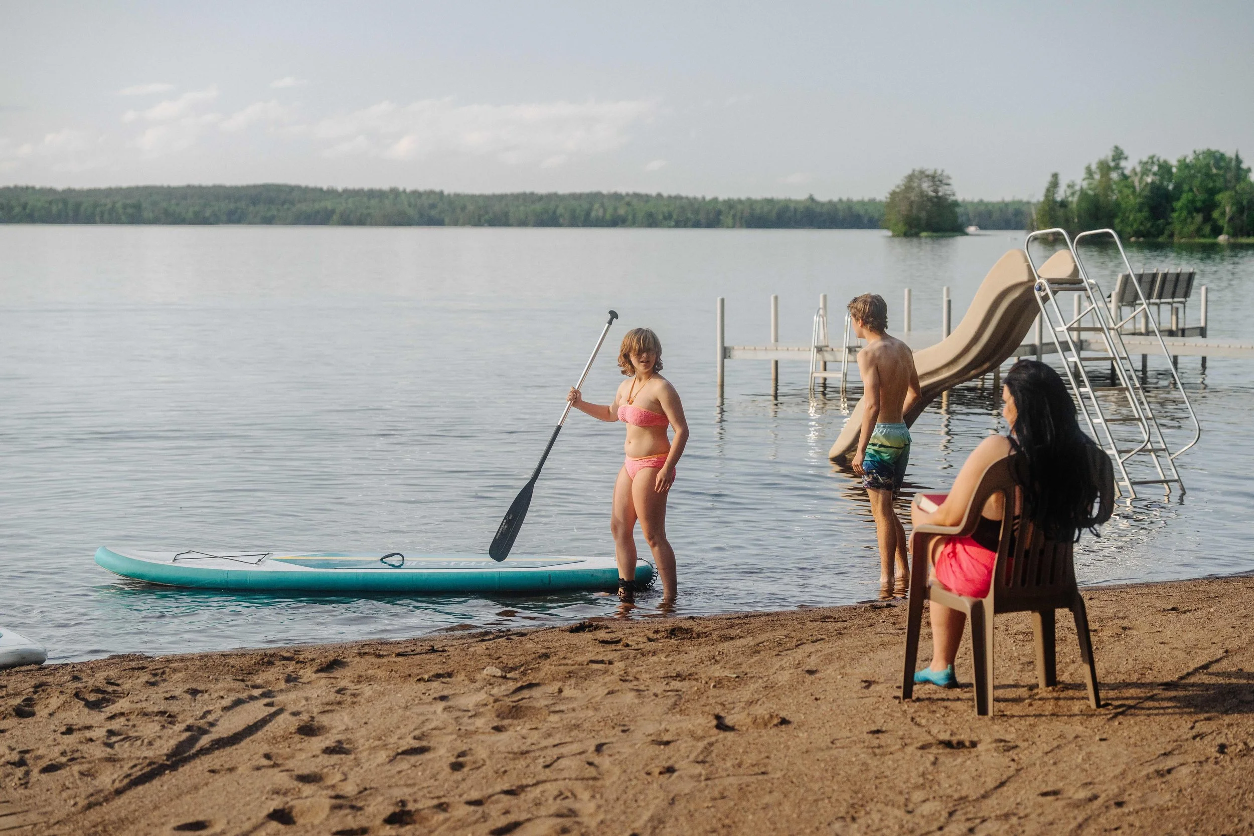 People at a lakeside, woman sitting on a chair reading, two kids by the water, one on a paddleboard, the other standing near a slide.