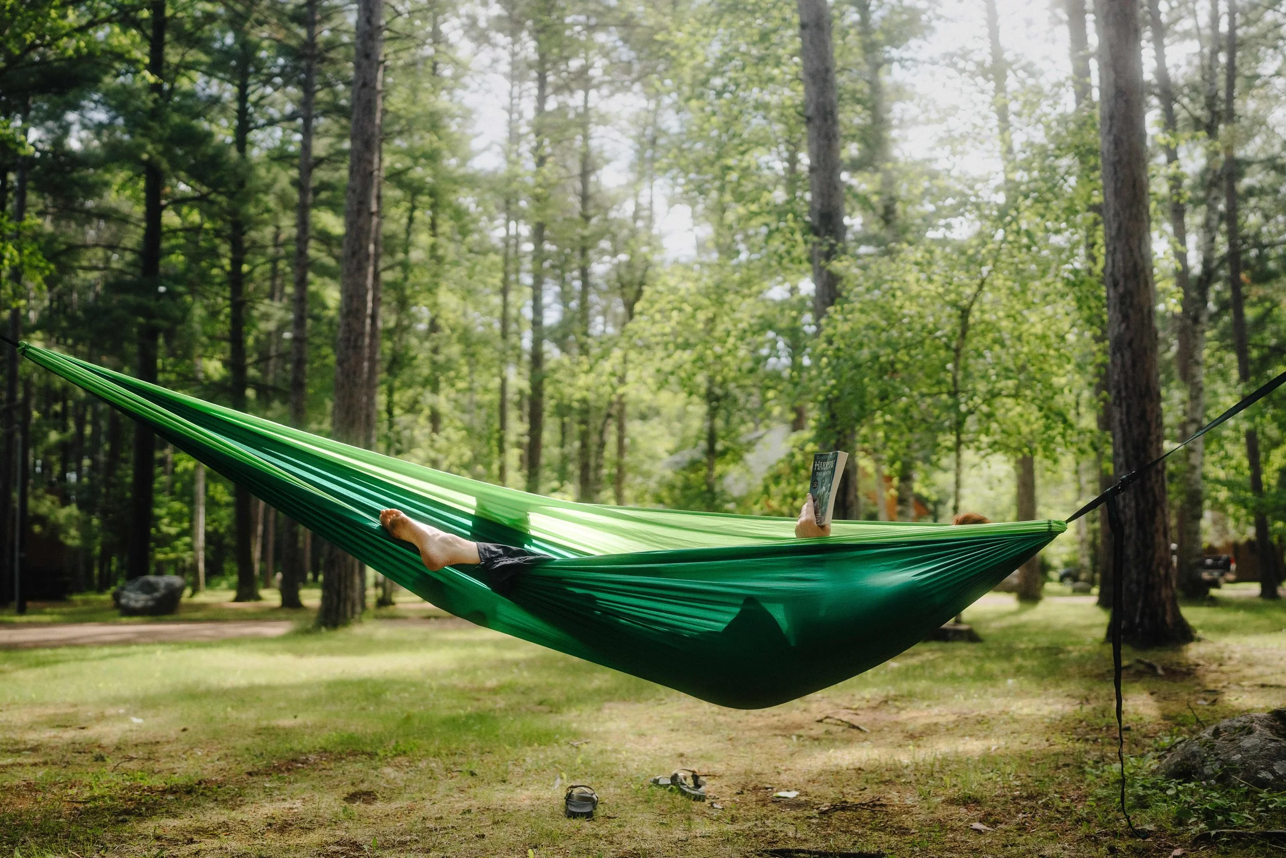Person lying in a green hammock reading a book in a wooded park.