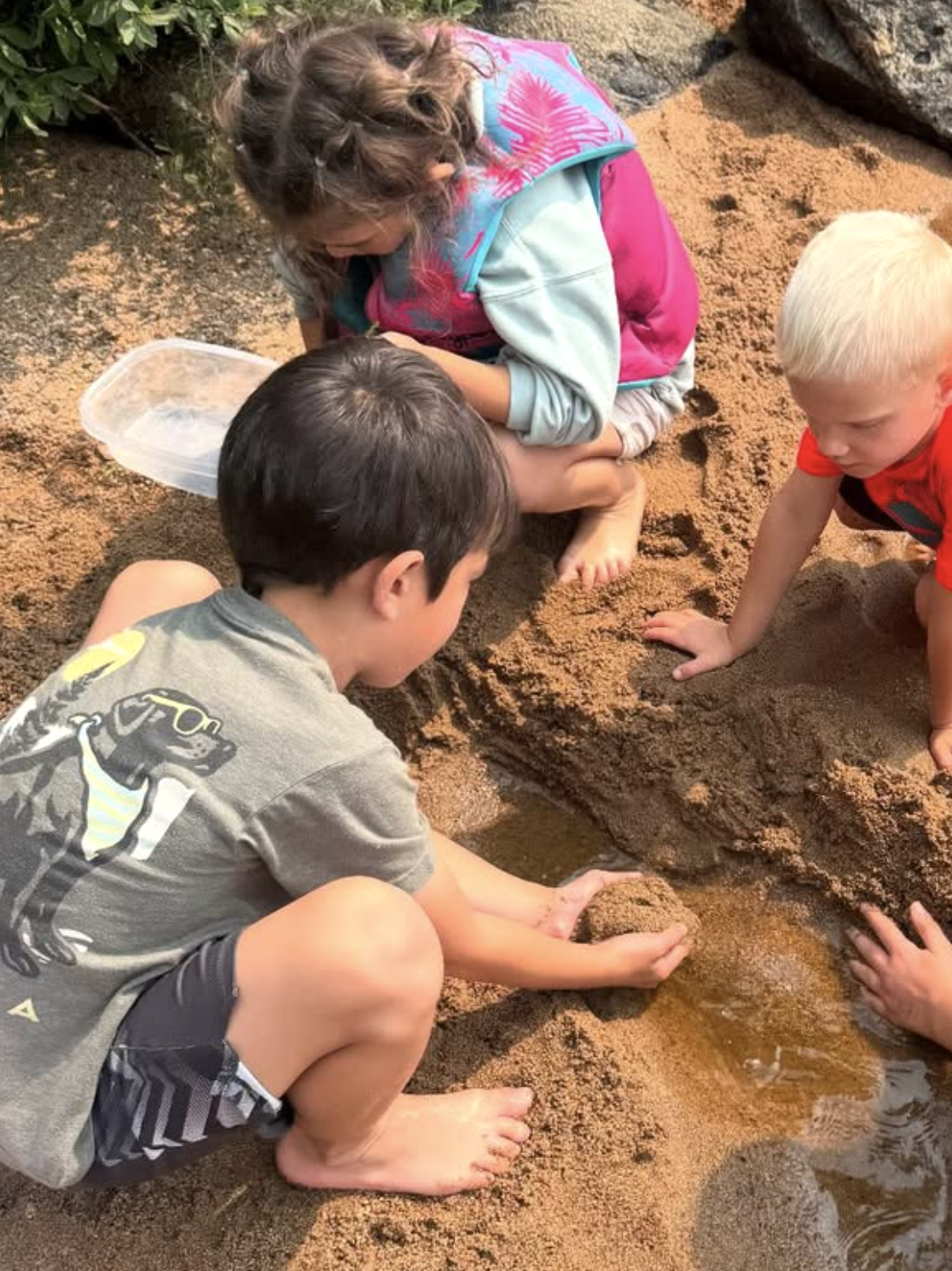Three children exploring and playing in a sandy creek bed, using their hands to dig and find water.