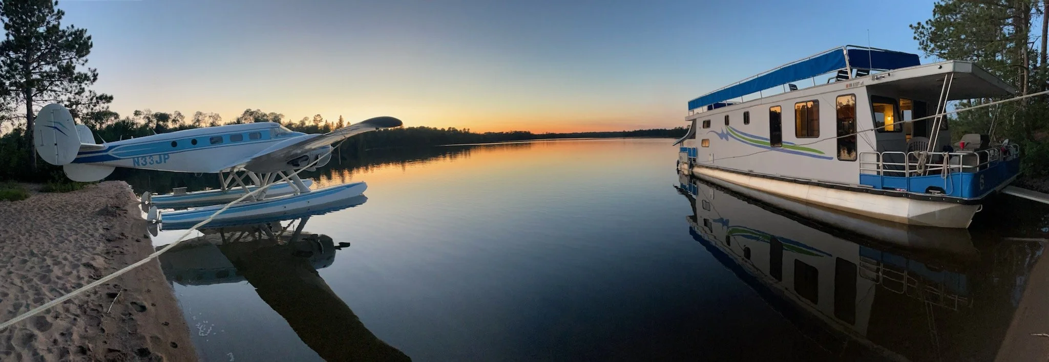 A seaplane and a houseboat docked on Birch Lake at sunset, with trees and a colorful sky reflected on the water.