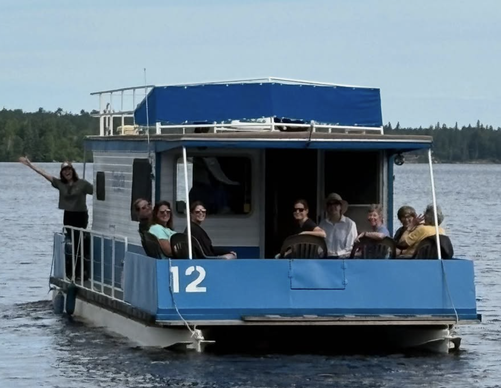 A group of people on a blue and white pontoon boat on a lake, some seated and some standing, with a person waving from the back of the boat.