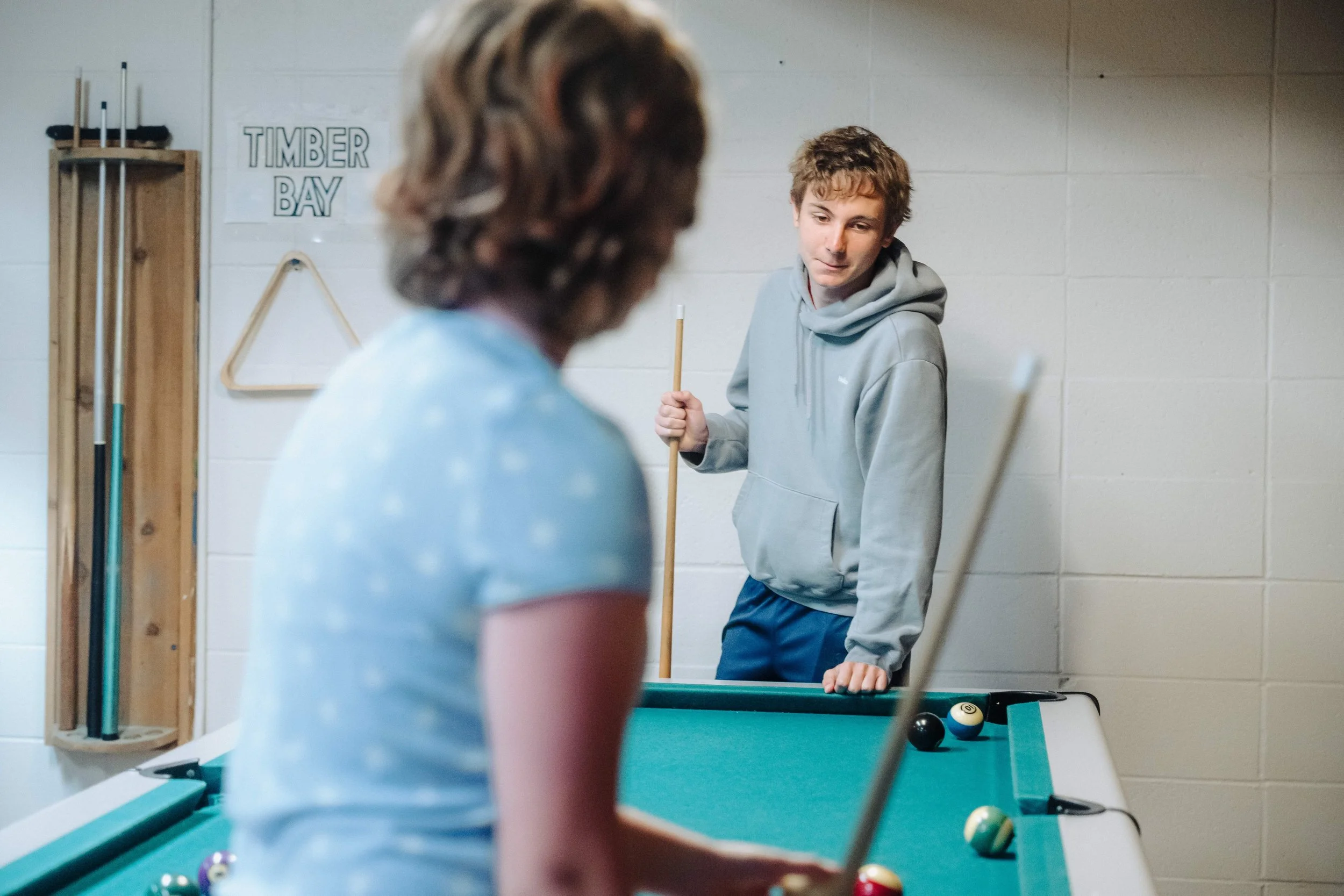 A young man and a woman are playing pool in a game room. The young man is standing by the pool table, holding a cue stick, and looking at the woman. The woman is positioned closer to the camera, preparing to take a shot. The game room has a cue rack on the wall labeled "Timber Bay" and a triangular fellow hanging on the wall.