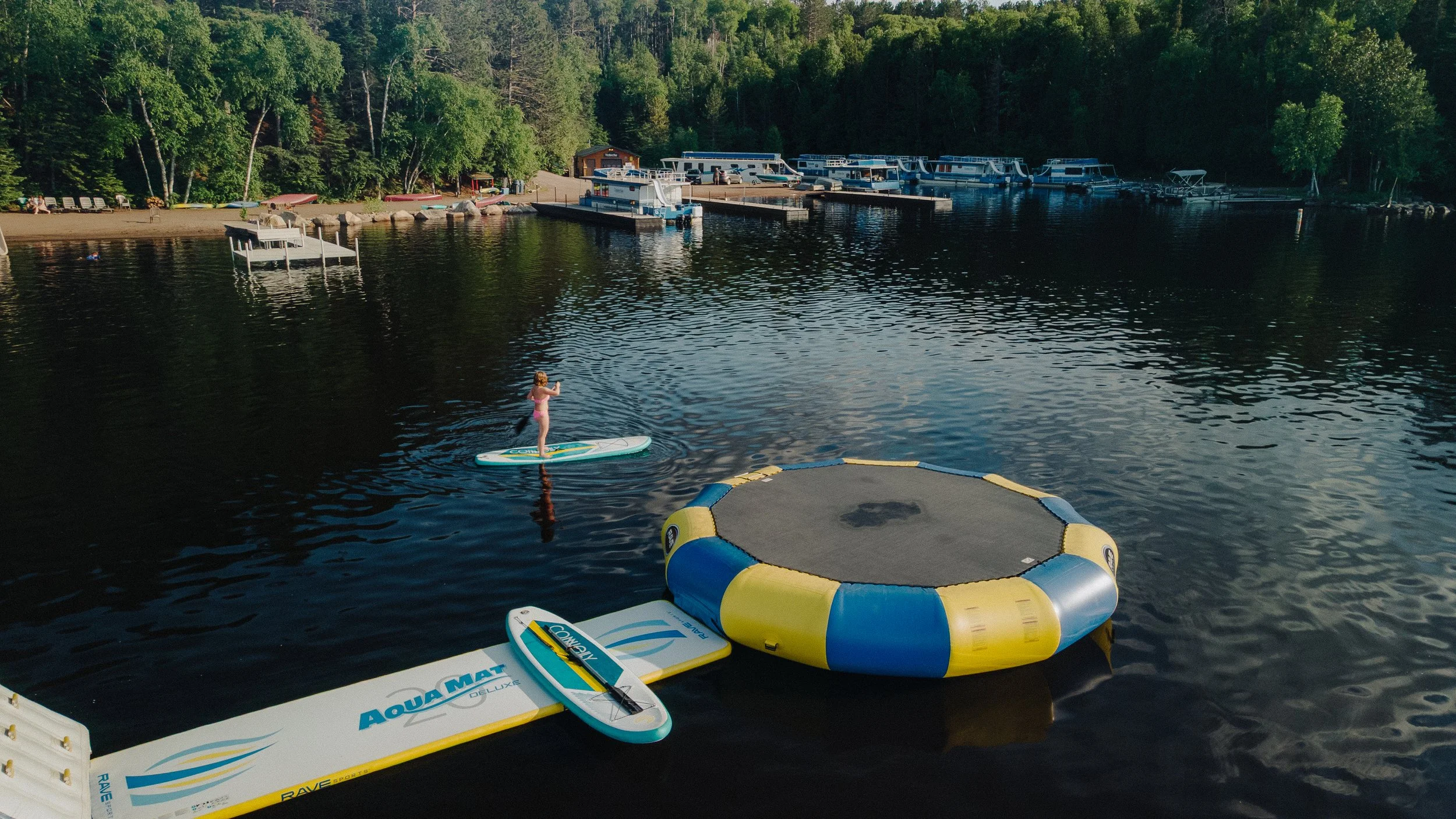 A person in a pink swimsuit is paddleboarding on a lake near a large, circular inflatable water trampoline, with paddleboards tied to the dock. The background shows a wooded shoreline with boats, docks, and a small building.