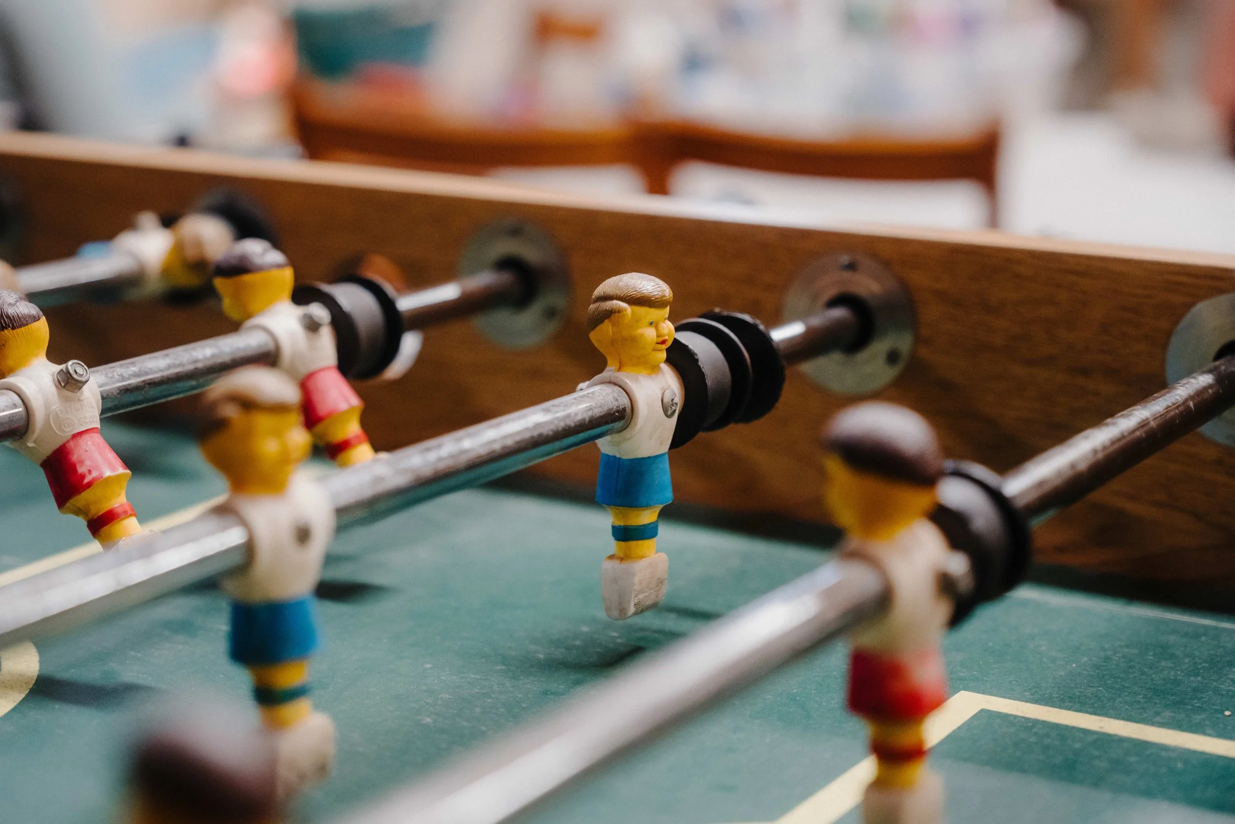 Close-up of a vintage foosball table with colorful player figures, featuring one in the center wearing a white shirt and blue shorts, with other players blurred in the background.