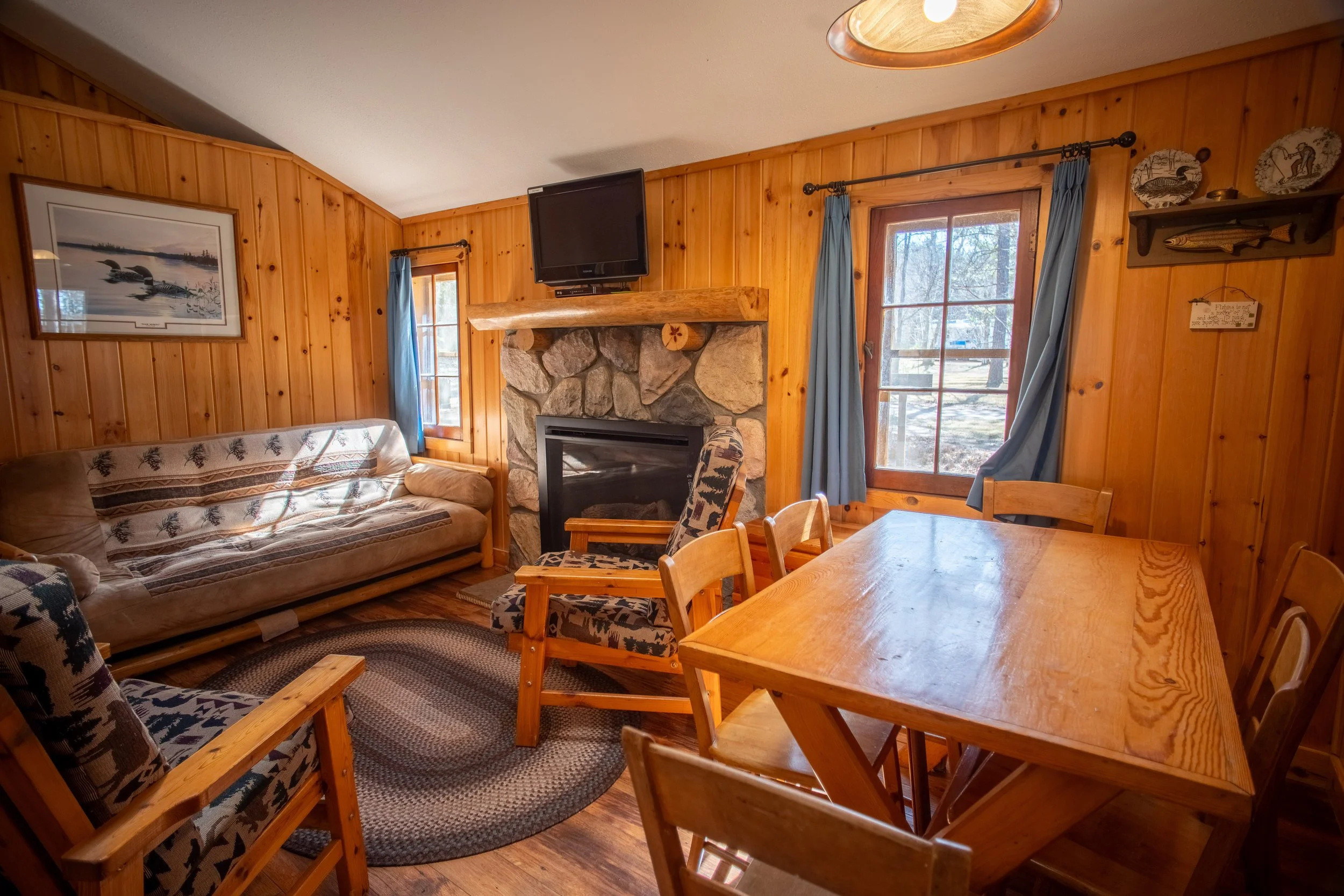 A cozy living room with wood-paneled walls, a stone fireplace with a mounted TV above, a wooden dining table with matching chairs, a sofa with a patterned cover, and decorative plates on the wall. Large windows with blue curtains show a snowy outdoor scene.