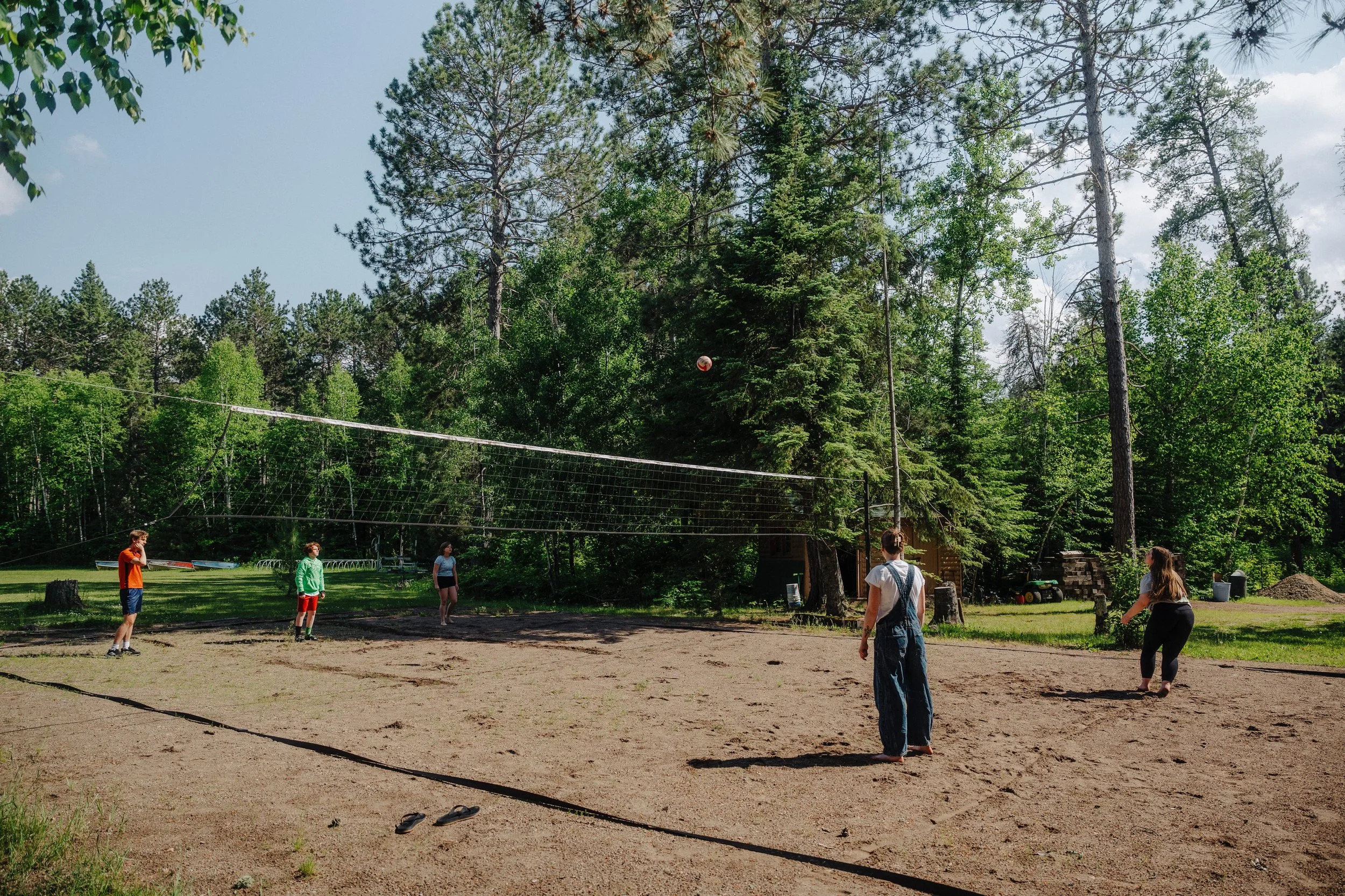 People playing volleyball on a sand court surrounded by green trees in a forested outdoor area.