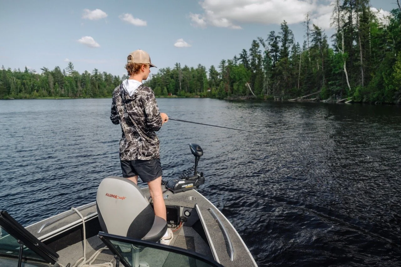 A young person fishing from a boat on a lake, surrounded by trees and a partly cloudy sky.