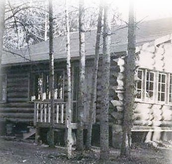 A log cabin with a small porch and multiple windows, surrounded by tall trees in a wooded area.