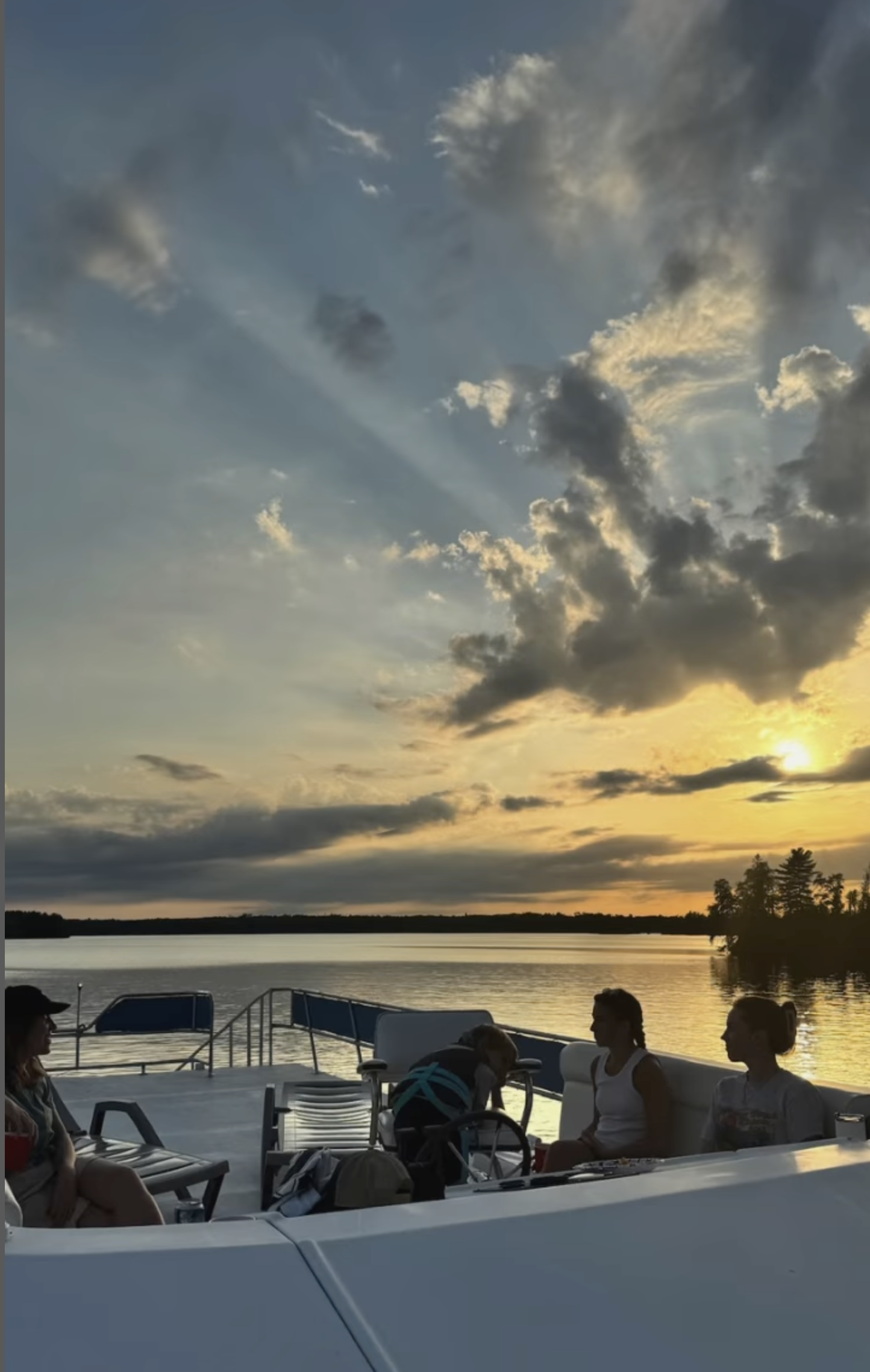 People sitting and relaxing on a boat during sunset over a lake with a cloudy sky and treeline in the background.