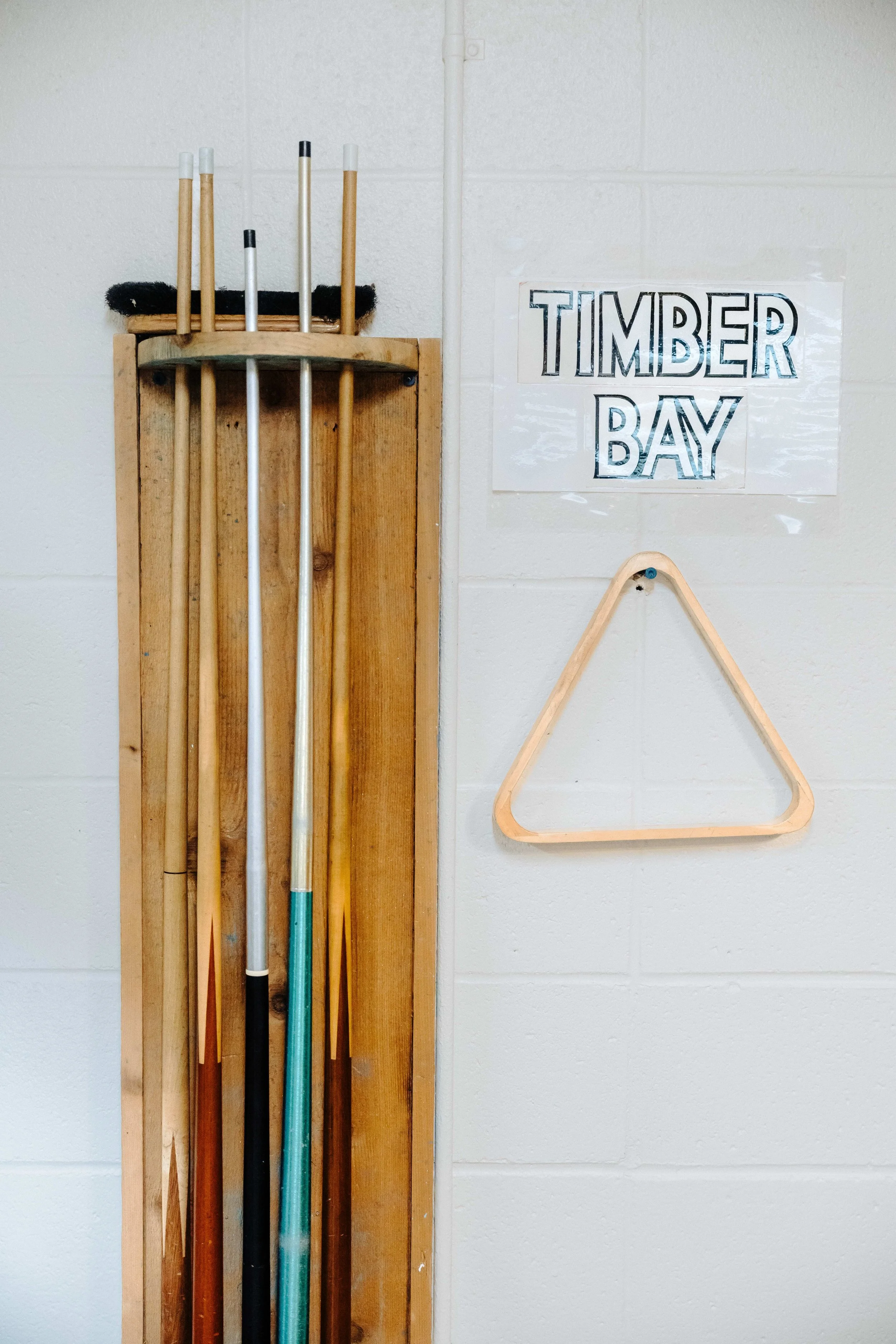 A wooden rack holding several pool cues, with a sign that reads 'Timber Bay' on a white brick wall.