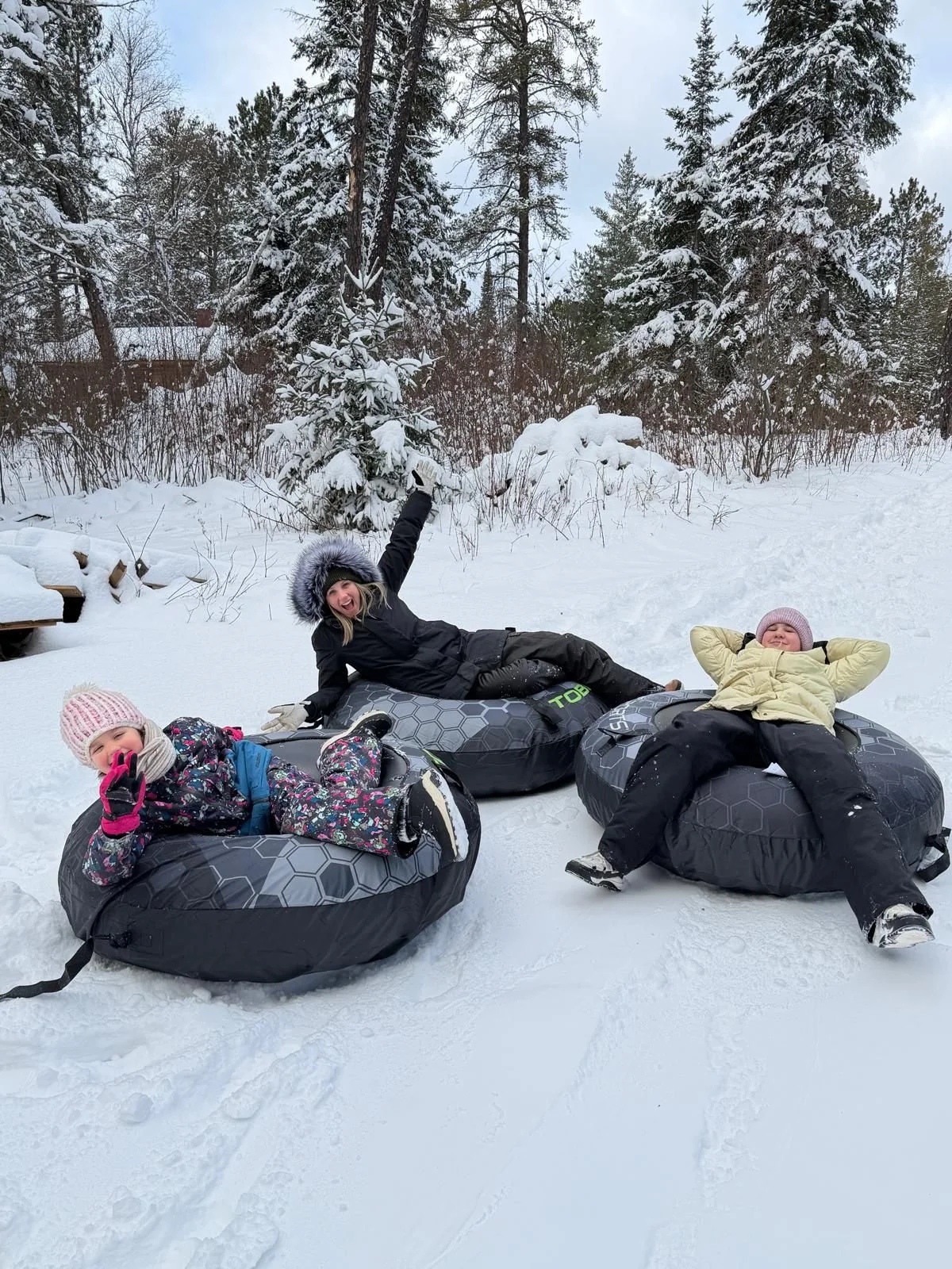 Three children in winter clothing relaxing on snow tubes in a snowy outdoor setting with snow-covered trees and bushes in the background.