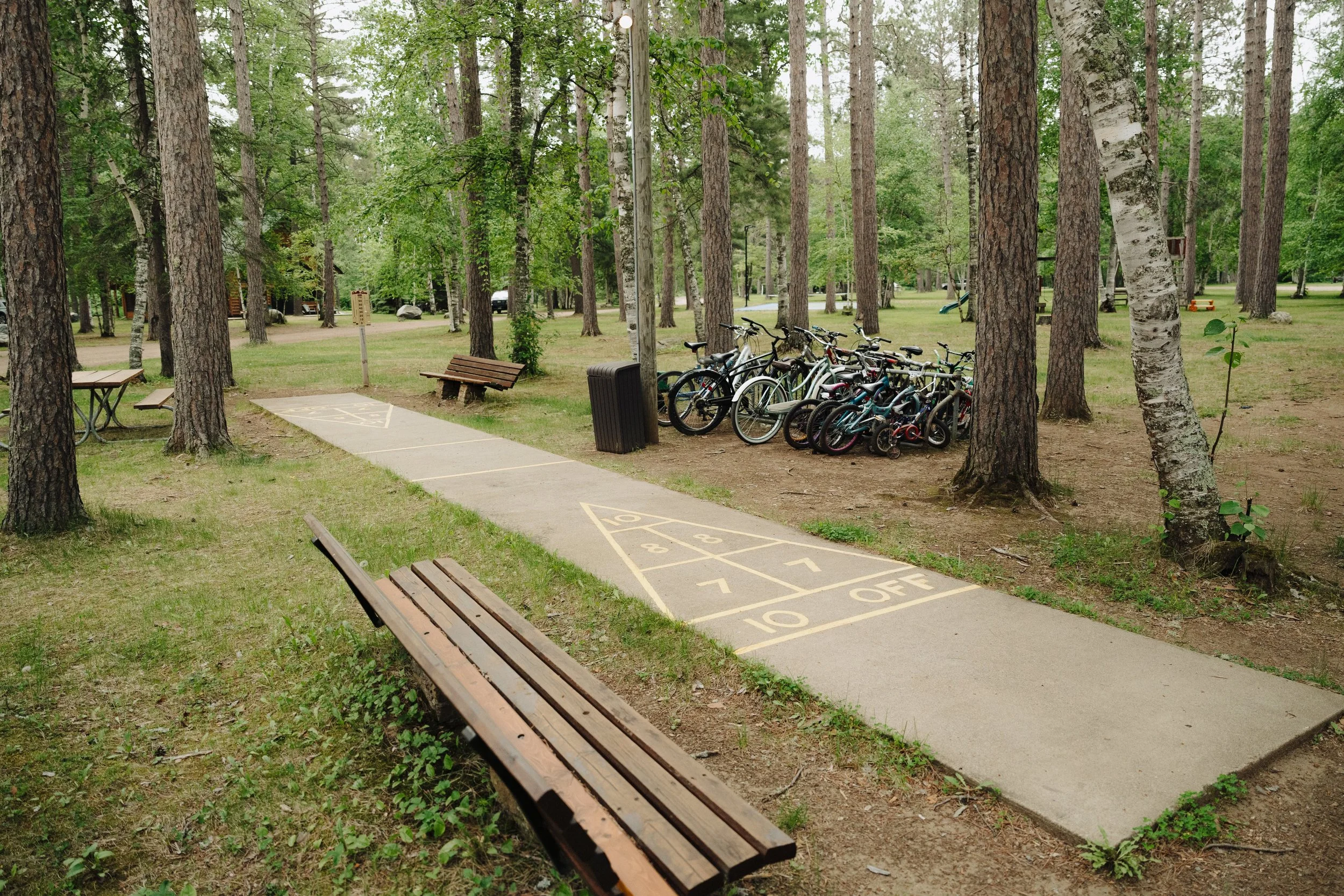 A park scene with a paved game of hopscotch on the sidewalk, benches, a trash can, and a row of bicycles parked among trees.