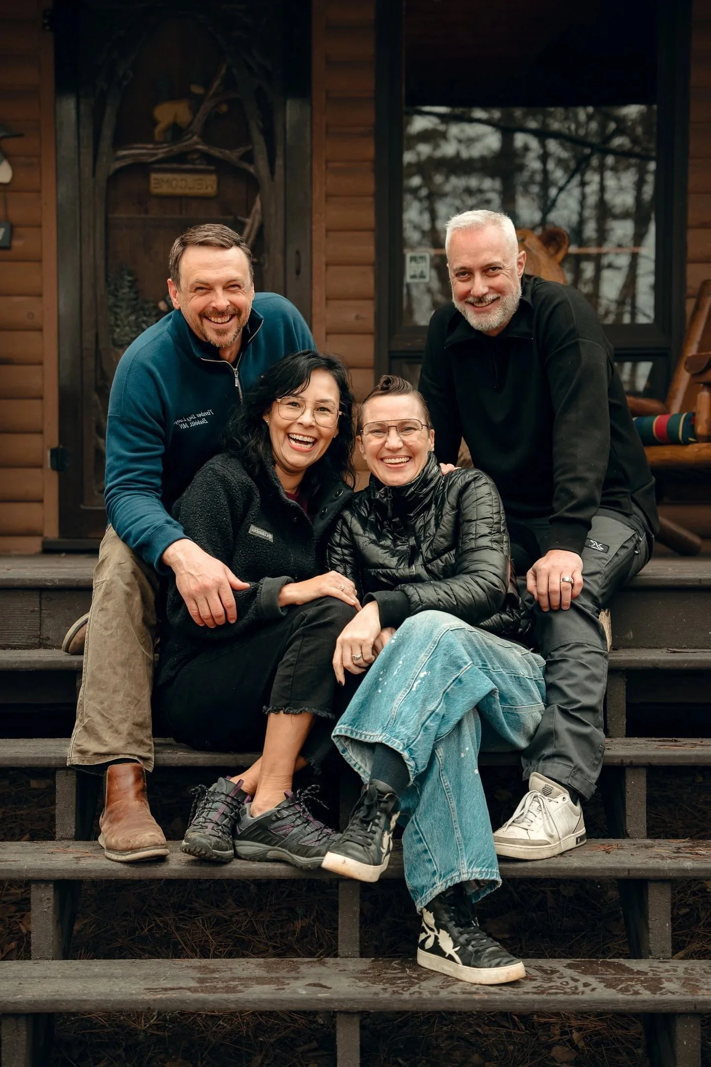 Four friends sitting on steps in front of a log cabin, smiling at the camera.